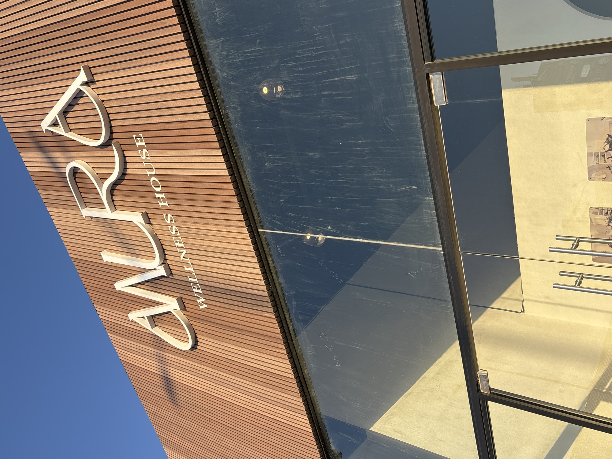 The image shows the facade of "Aura Wellness House" in San Bernardino Tlaxcalancingo, Mexico. The facade features horizontal wooden paneling with the name "AURA WELLNESS HOUSE" in white lettering. A glass entrance with a dark frame is visible to the right, reflecting the sky and surrounding environment. Metal handles are visible on the interior side of the glass door. The sky is visible in the lower left corner.