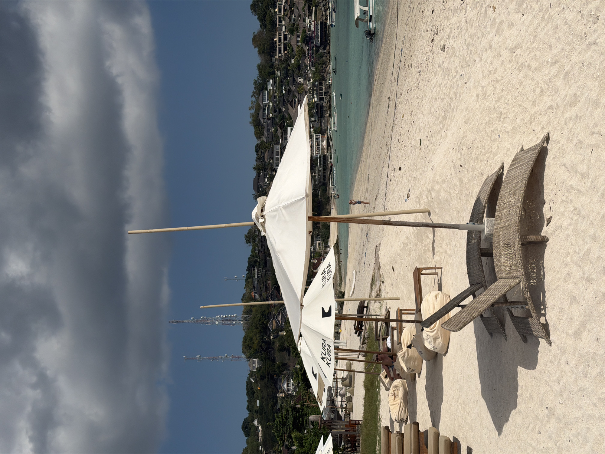 The image depicts a serene beach scene in Nusa Penida, Indonesia. In the foreground, two woven lounge chairs are positioned on the sandy shore, casting long shadows. Behind them, several large white umbrellas are set up, some bearing the "KURA KURA" logo. Scattered around the umbrellas are numerous beanbag chairs. A person is visible in the middle ground, standing on the beach and looking out towards the water, dressed in a blue top and denim shorts.
The ocean in the background is a clear turquoise, dotted with several small boats. The coastline is lined with lush green vegetation and a cluster of buildings, suggesting a developed resort area. In the far distance, on the hilly terrain overlooking the coast, are tall communication towers, characteristic of areas with extensive infrastructure. The sky above is a dramatic mix of dark, heavy clouds and patches of clear blue. The overall atmosphere is one of a tranquil, albeit slightly overcast, day at a tropical beach.