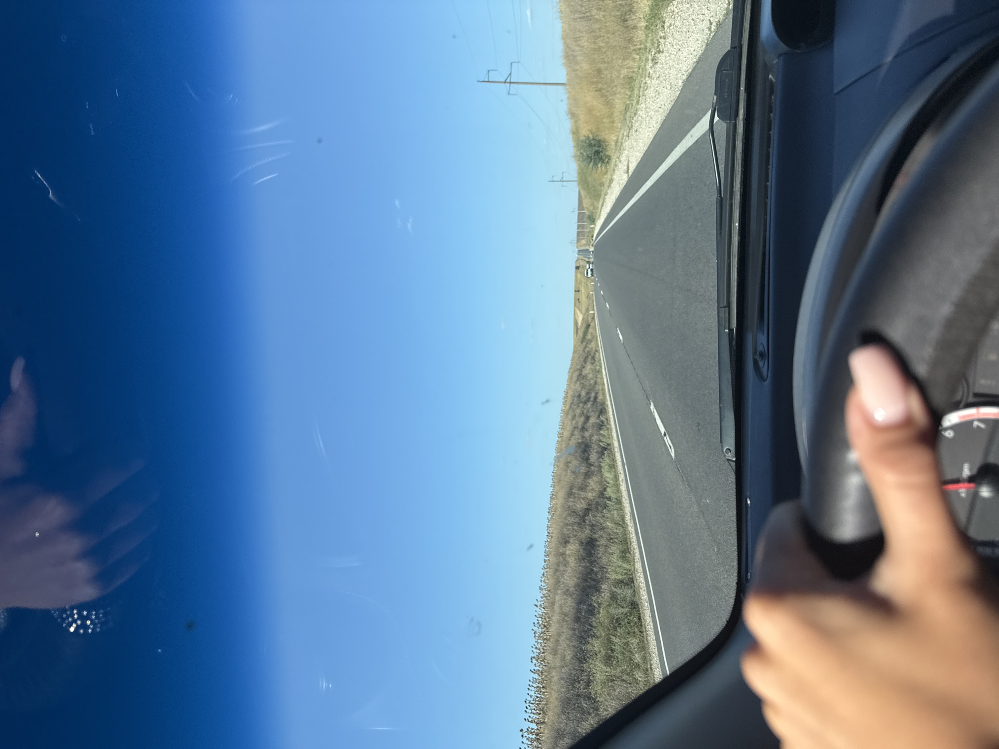 A first-person perspective shot from the driver's seat of a vehicle shows a hand on the steering wheel. The road ahead is asphalt with a white line marking the lane. The surrounding landscape is dry and grassy, with a line of tall, dried plants on the left side. In the distance, there are power lines and a few other vehicles. The sky is clear and blue. The image is likely taken on a sunny day. Based on the visible power pole design and surrounding vegetation, the location is presumed to be Comrat, Moldova.