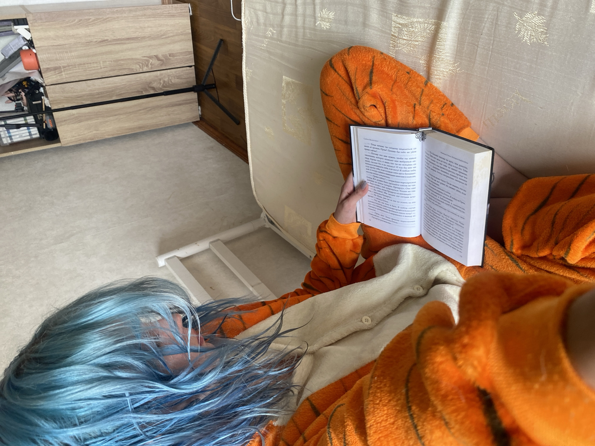 In a sunlit room, a person with vibrant blue hair lies on a bed, engrossed in a book. They are wearing a plush orange onesie with black tiger stripes, adding a playful touch to the scene. The book, held open in their hands, displays Russian text. Beside the bed, a light-colored wooden dresser is visible, partially filled with various items. A stark white bed frame is also present. The flooring is a light-colored carpet. The overall impression is one of quiet relaxation and a moment of personal escape. The lighting suggests it is daytime.