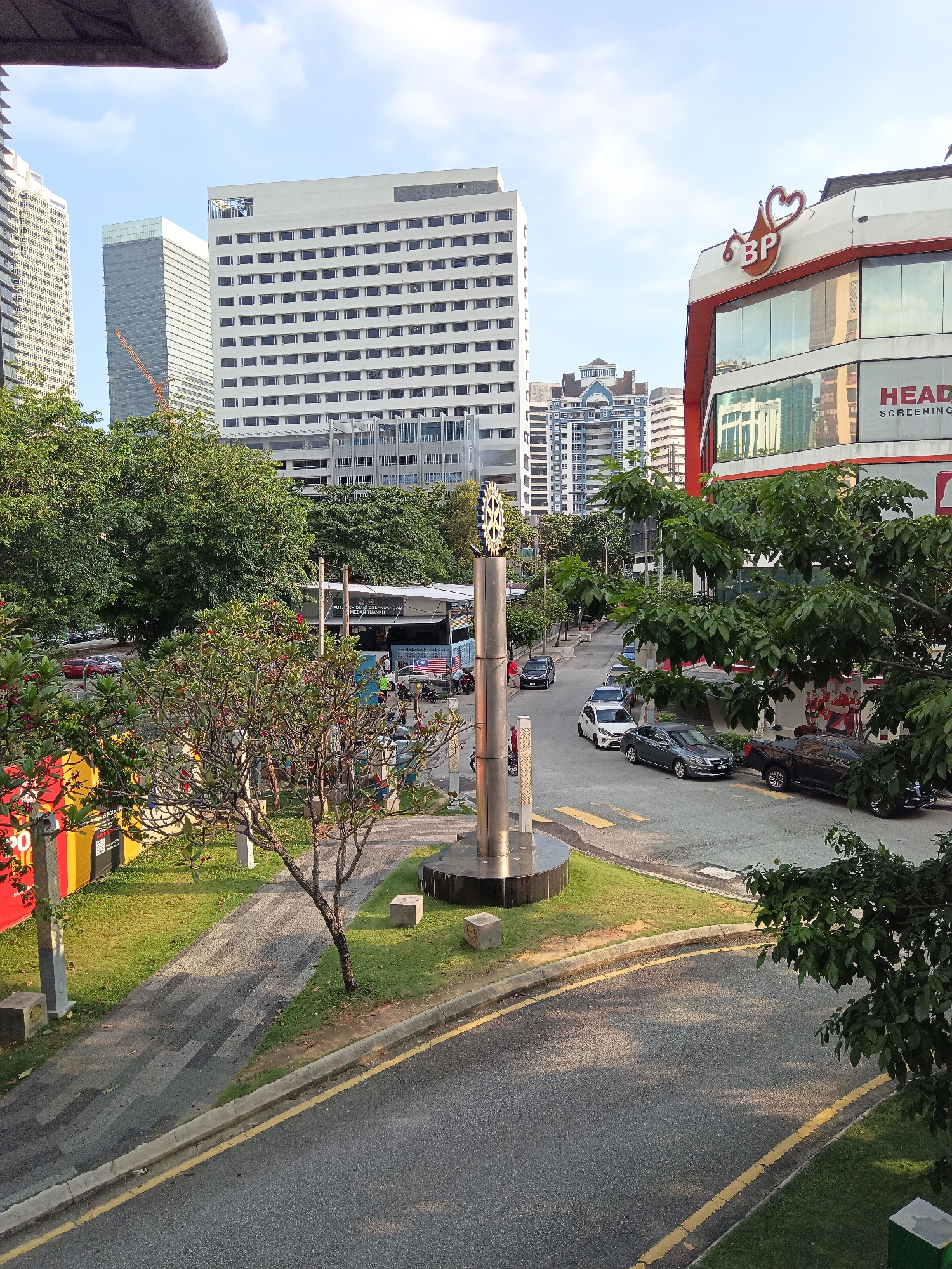 The image depicts an outdoor urban scene in Kuala Lumpur, Malaysia, with a focus on a street intersection and surrounding buildings. In the foreground, a paved road curves to the right, bordered by a grassy median and a sidewalk. A cylindrical monument with a gear-like structure at the top stands in the median, accompanied by a small, bare tree. Several cars, including a black SUV, a grey sedan, a white hatchback, and other vehicles, are visible on the road.
In the mid-ground, lush green trees line the street, partially obscuring a covered walkway or structure with signage, including a Malaysian flag. Behind the trees, several multi-story buildings rise, including a large white building with numerous windows and a modern facade. To the right, a building with a red and white exterior displays the "BP" logo prominently and signage indicating "HEAD SCREENING." Further in the background, more tall buildings, characteristic of a city skyline, are visible under a partly cloudy sky. The lighting suggests it is either early morning or late afternoon, with sunlight casting shadows on the road. A construction crane is visible behind one of the buildings. The overall impression is of a well-developed urban environment with a mix of commercial and residential structures.