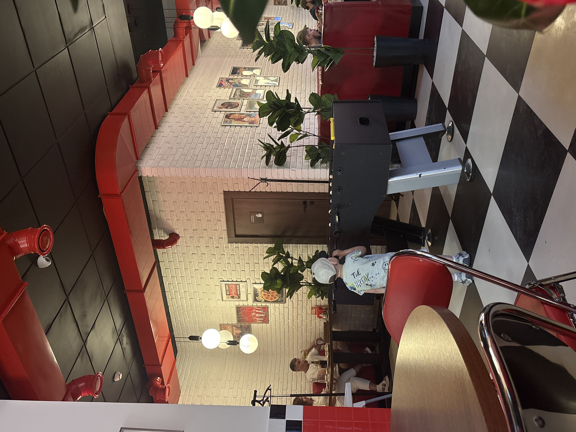 The image captures an interior scene, likely of a restaurant or diner, with an American-retro theme. The location is identified as Perm, Russia. The ceiling features black tiles and red ductwork. The walls are white brick adorned with framed images. The flooring is black and white checkered. A foosball table is positioned in the center-right of the image.
A young boy wearing a white cap and a blue t-shirt stands near the foosball table. Further back in the image, seated at tables, are what appear to be customers: one man, and two women. A table and diner style chairs are located in the foreground, in front of them a small child.
The overall scene conveys an atmosphere of a casual dining establishment with some elements of entertainment.