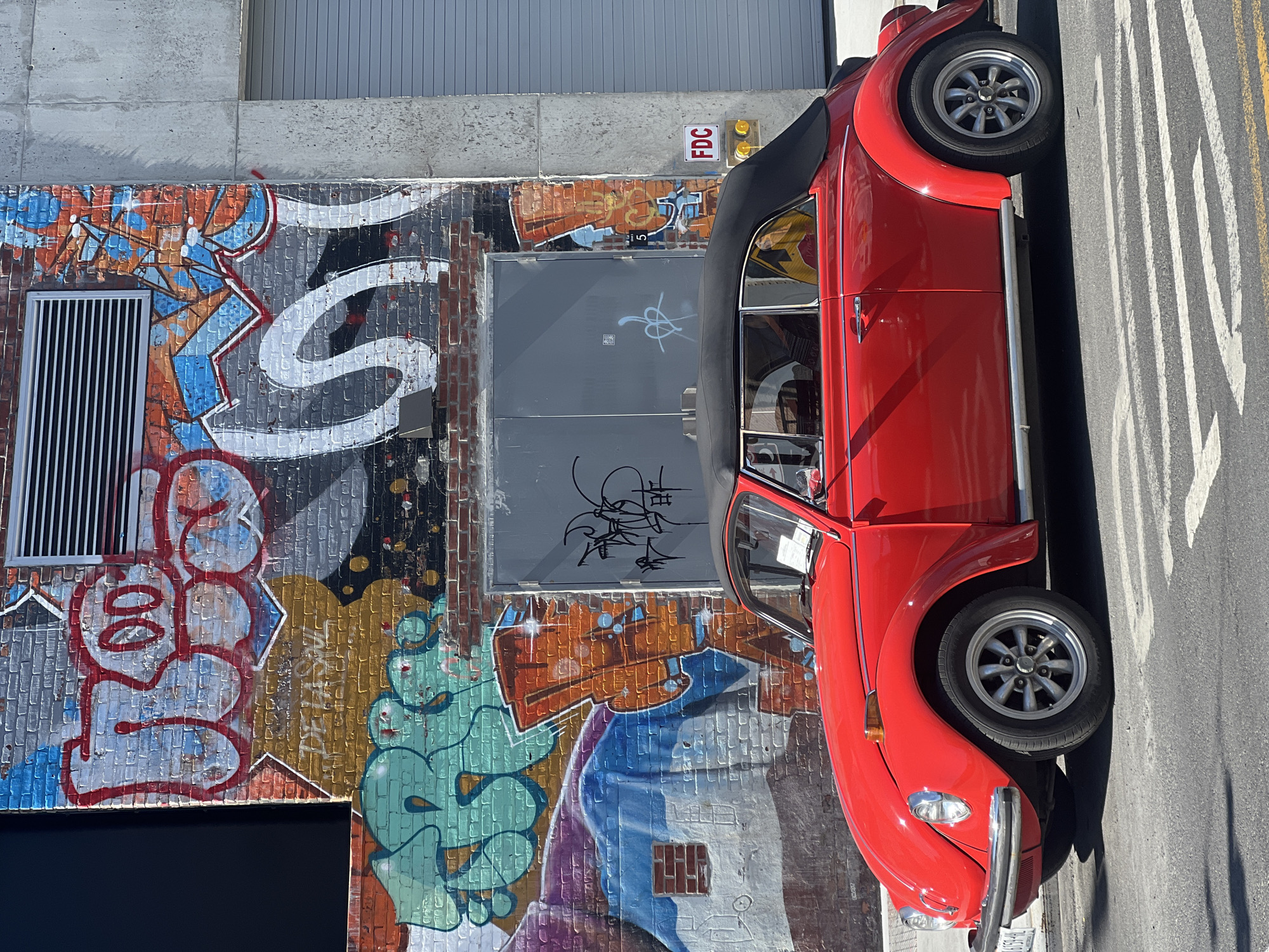 A red Volkswagen Beetle convertible is parked on a city street next to a brick building covered in graffiti. The building has a gray metal door with more graffiti and an FDC sign above it. The street is marked with parallel white lines. The location appears to be in an urban area, possibly New York City.