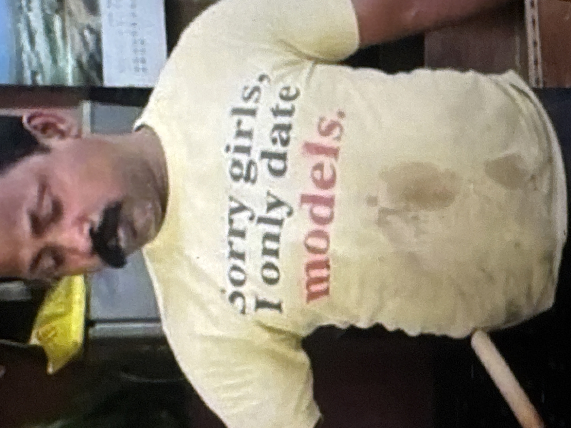 In Kuta Selatan, Indonesia, a man wearing a light yellow t-shirt with the text "sorry girls, I only date models." printed in gray and red stands against a blurred background. The t-shirt has noticeable sweat stains on the front. To the left of the man, a blurred object resembling a wooden stick is visible. The man has a dark mustache and appears to be looking downwards. The environment is dimly lit, with hints of shelving or equipment in the background.