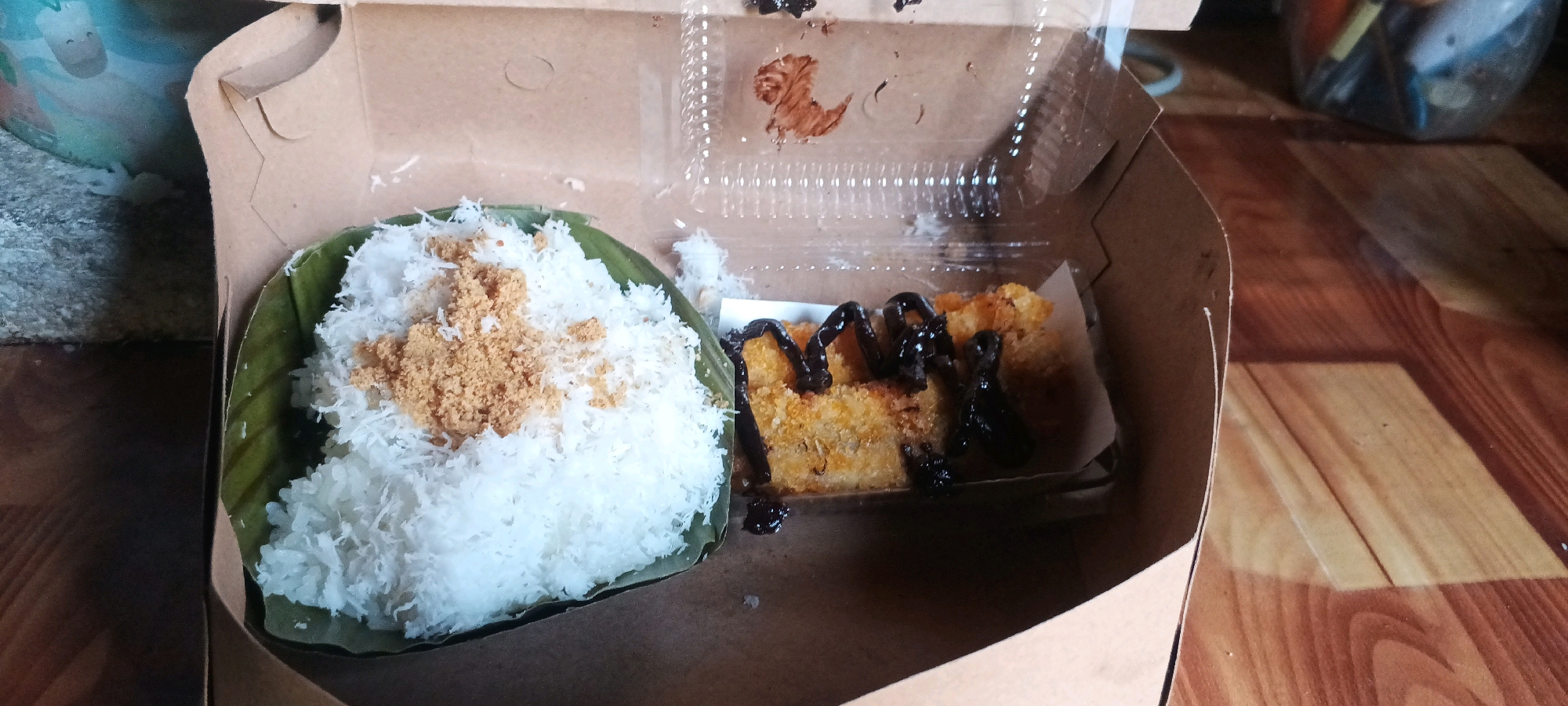 In a brown cardboard box, two food items are presented. On the left, a mound of white rice, possibly sticky rice, is served on a green banana leaf and topped with a scattering of brown sugar. To the right, a rectangular piece of fried food, possibly a snack like pisang goreng (fried banana), is drizzled with dark chocolate sauce. The box is open, revealing a transparent plastic lid inside. The background shows a patterned wooden surface. The image was taken in a residential setting in Penanggungan, Indonesia.