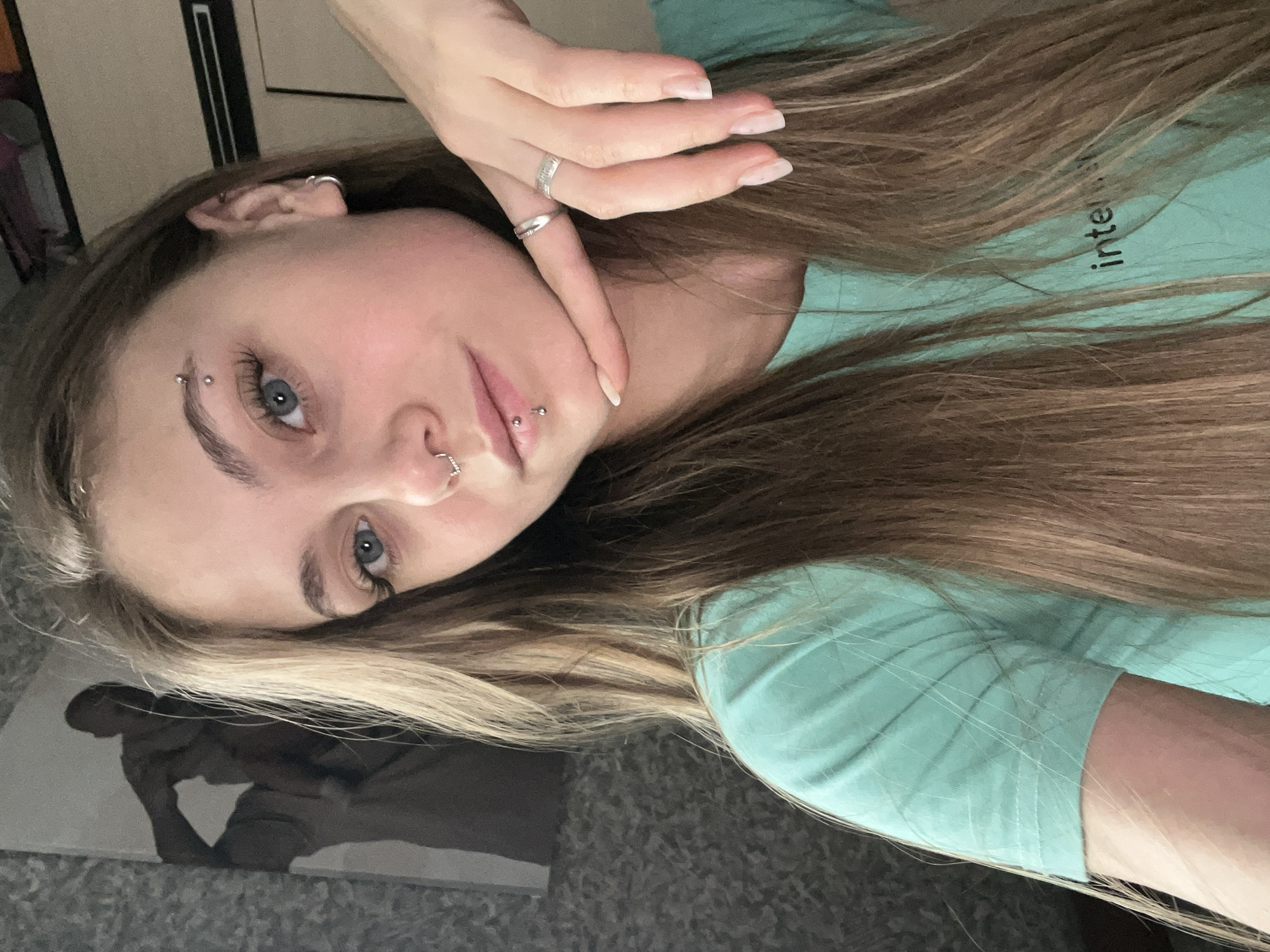 A young woman is looking at the camera. She has light blue eyes, fair skin, and long, light brown hair. She is wearing a turquoise t-shirt and has multiple facial piercings including an eyebrow piercing, a nose ring, and lip piercings. She is wearing silver rings on her hand, which is placed near her face. The background is blurry, revealing a carpet, a partial image of artwork, and an undefined dark object. The city of Фоки, Russia, cannot be determined from the image.