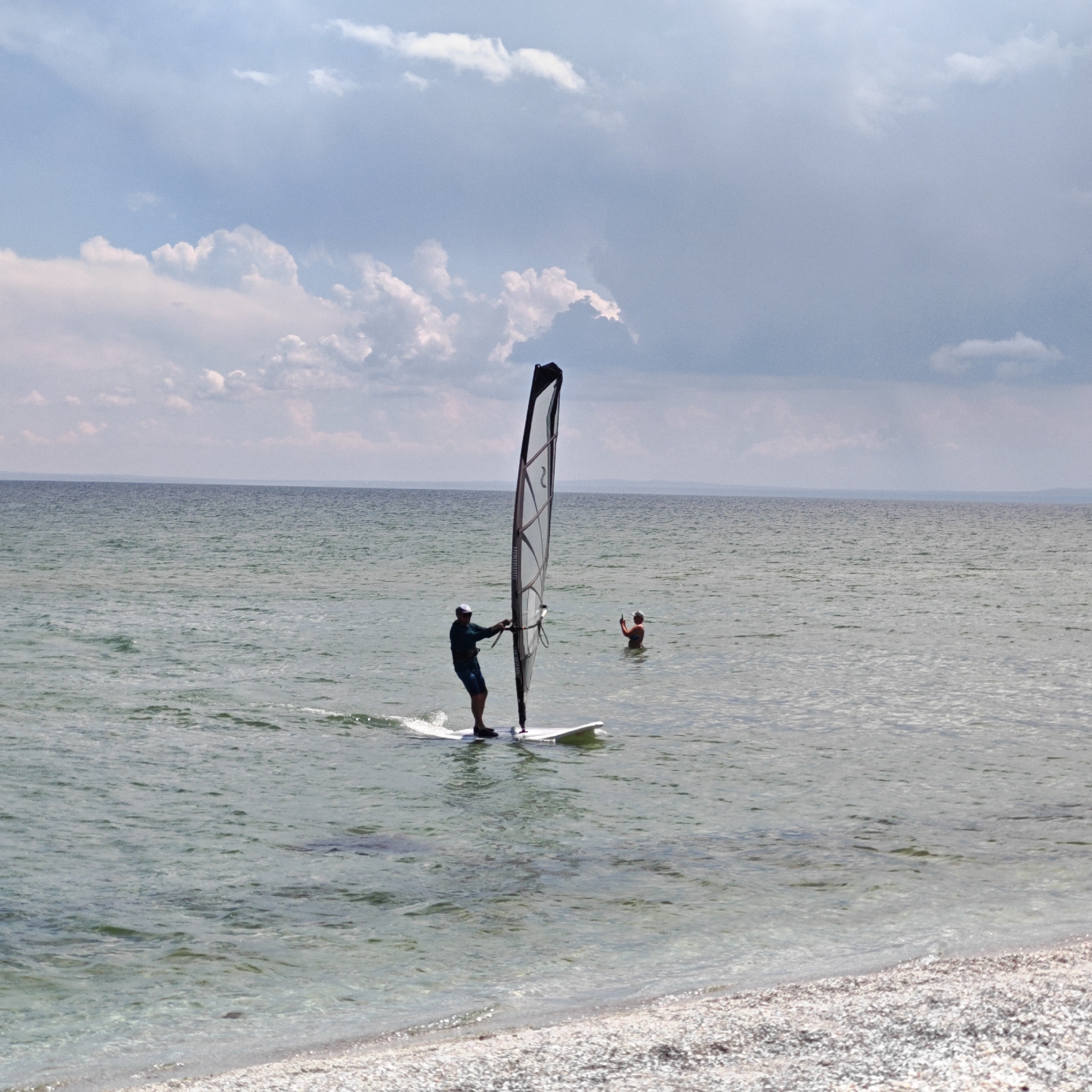 The image shows a serene seascape with a person windsurfing on a calm body of water. The sky is partly cloudy, with the sun casting a gentle light. In the foreground, a beach is visible, covered with small pebbles and shells. The windsurfer, silhouetted against the bright sky, is positioned in the mid-ground, actively engaged in the sport. Another person is visible in the water further back, seemingly observing or participating in a different activity. The overall impression is one of peaceful outdoor recreation in a coastal environment.