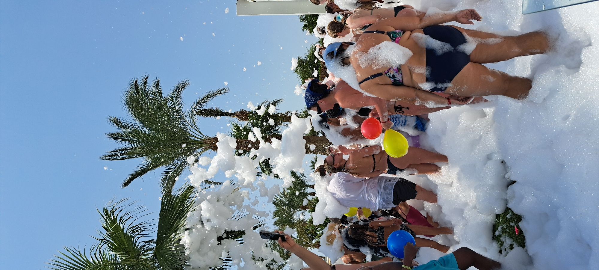 This is a vertical, eye-level, medium-shot photograph taken outdoors in Sharm El Sheikh, Egypt. The scene depicts a foam party taking place among palm trees under a clear blue sky. A large quantity of white foam fills the foreground and mid-ground, immersing a group of people, most of whom are wearing swimwear. Several individuals are holding colorful balloons, including a blue one in the left mid-ground, a yellow one in the center mid-ground, and a red one in the center mid-ground. Palm trees are visible in the background. The overall atmosphere appears to be festive and energetic.