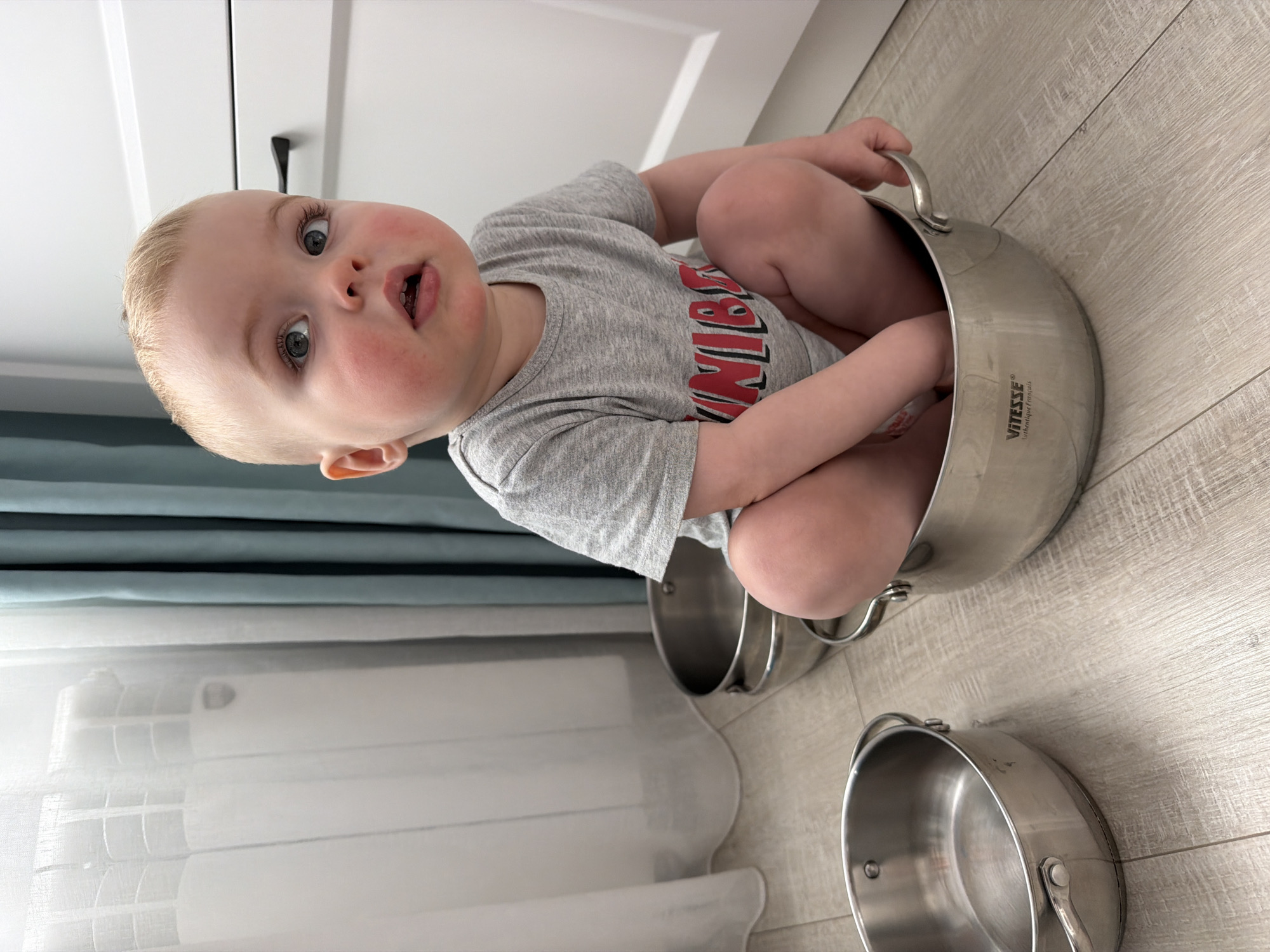 A young child is sitting inside a stainless steel cooking pot. Two other cooking pots are on the floor near the first pot. The floor is light-colored wood plank. The child wears a gray t-shirt with red text. In the background are white cabinets, a curtain, and a window. The cooking pot brand is "Vitesse La Boutique Français." The image was not taken in Votkinsk, Russia.