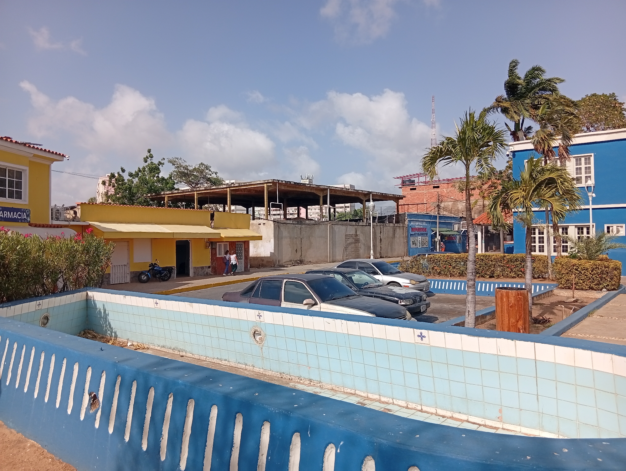 The image captures a sunny day in Juan Griego, Venezuela. The scene features a tiled, empty, blue and white fountain in the foreground, with an open design on its blue exterior. Behind the fountain are parked cars. Further in the background are buildings of different colors, including a yellow structure with a motorcycle parked in front, a blue building with white windows, and a partially constructed building with a skeleton structure. Palm trees are situated near the blue building. A few individuals are present in the background, one standing in the doorway of the yellow building. The sky is blue with scattered white clouds.