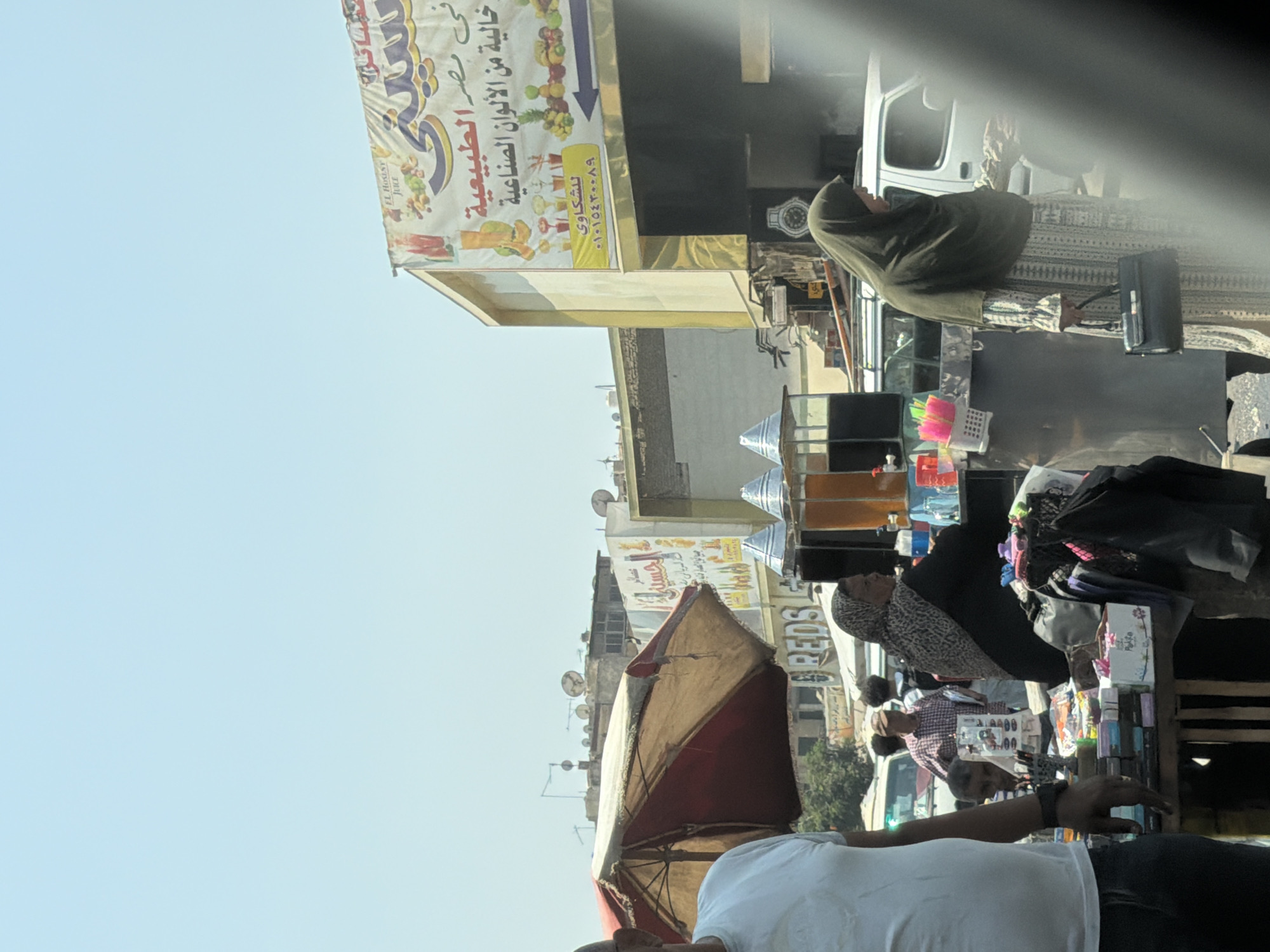 The image captures a bustling street scene in Cairo, Egypt, likely during daylight hours given the bright sunlight and clear sky. In the foreground, a person wearing a white t-shirt and dark pants stands with their left arm bent, appearing to gesture or hold something out of frame. Further back, a woman in a patterned headscarf and dark clothing stands behind a stall displaying various items, possibly for sale. To her right, another woman in a dark green hijab and a patterned dress walks past, carrying a black handbag.
The background reveals a commercial area with shops and stalls. A prominent banner with Arabic script and images of fruits and drinks hangs on a building. Nearby, a red and beige umbrella shades a small seating area. Several vehicles, including a white van, are visible on the street, suggesting it's a busy thoroughfare. The architecture appears typical of urban Egypt, with buildings of varying heights and satellite dishes on rooftops. The overall impression is one of daily life and commerce in a lively city environment.
