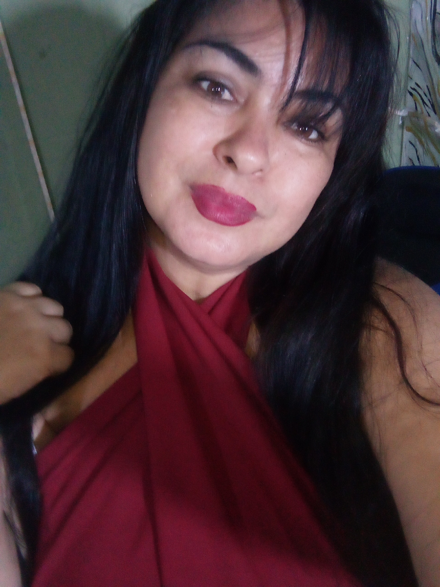 A woman is shown in a medium close-up shot. She has dark hair with bangs, and brown eyes. She is wearing a dark red top that crosses over her chest. The background appears to be an interior setting. There is a wall visible on the left side and a patterned surface on the right. Without additional information, it is not possible to verify the location in Juan Griego, Venezuela.