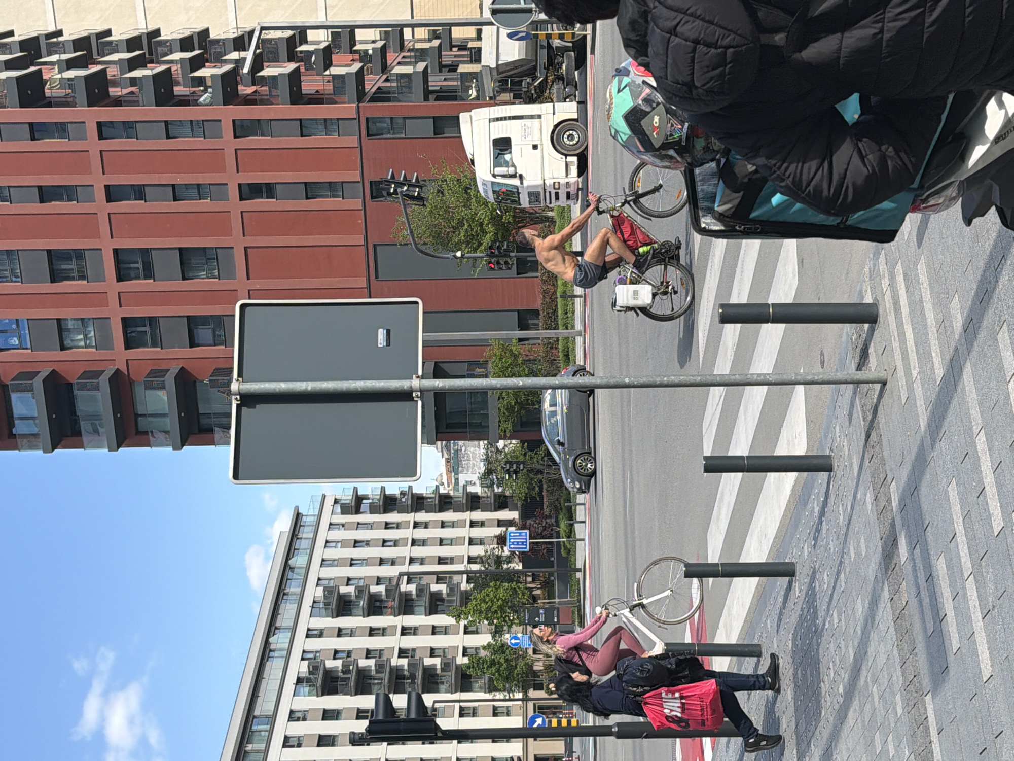 This image captures a vibrant urban street scene in Belgrade, Serbia, on a bright, sunny day with a clear blue sky and a few scattered clouds. The street is lined with modern buildings, including a prominent reddish-brown building with numerous rooftop units and a lighter-colored building featuring balconies.
In the center of the frame, a shirtless man with a beard, wearing shorts, performs a balancing act or wheelie on his bicycle. The bicycle is equipped with what appears to be a delivery box on its rear rack. Behind him, a white semi-truck is visible on the street, and a set of traffic lights hangs overhead.
On the right side of the image, on the sidewalk, a woman in pink athletic wear (leggings and a long-sleeved top) poses while holding a white bicycle, smiling directly at the camera. Closer to the foreground, partially obscured, are two other individuals. One wears a black puffer jacket and is next to a bicycle wheel, while the other, wearing dark clothing, carries a bright red shopping bag with the word "SALE" clearly visible.
The street features a zebra crosswalk, and several dark bollards line the edge of the patterned sidewalk. A large, blank grey sign stands prominently in the mid-ground. A dark car is also visible in the background behind the female cyclist. The scene suggests a busy but relaxed urban environment, capturing various activities on a typical day.