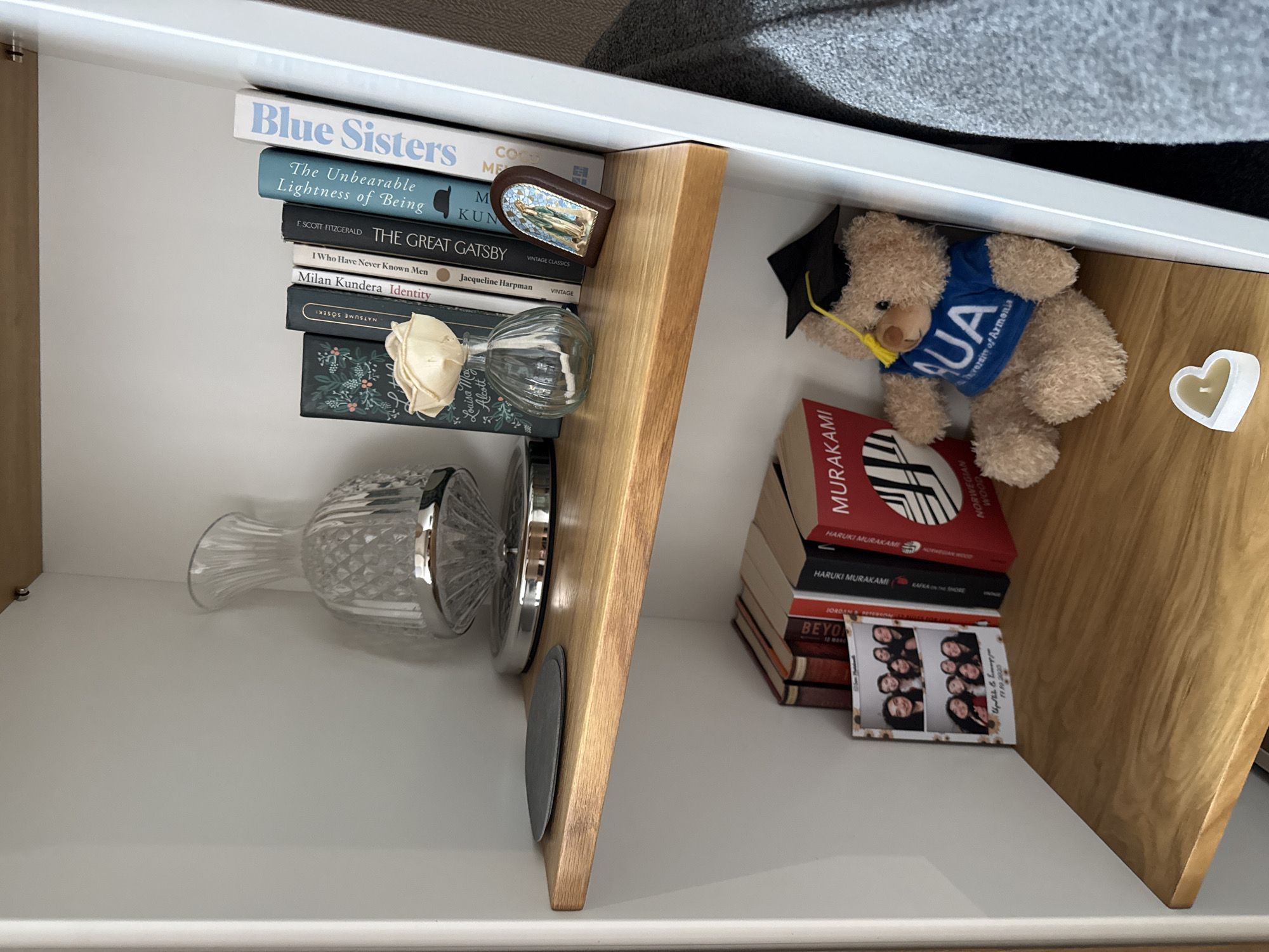 The image shows the interior of a shelf or drawer unit, likely within a domestic setting. On the left side, a stack of books is placed horizontally. The titles visible include "Blue Sisters," "The Unbearable Lightness of Being," "The Great Gatsby," "I Who Have Never Known Men," and "Identity." Above the book stack, there is a small, framed artwork. Beneath the books, there are two clear glass vases of different designs. On the right side of the shelf, there's a teddy bear wearing a graduation cap and a blue shirt with "AUA University of Armenia" printed on it. The books stacked next to the bear include works by Murakami. A small, framed photo sits next to the books. A heart-shaped candle holder is located on the right. The overall impression is of a collection of personal items. Given that the teddy bear is wearing a shirt for the American University of Armenia, this drawer is likely somewhere in Yerevan, Armenia.