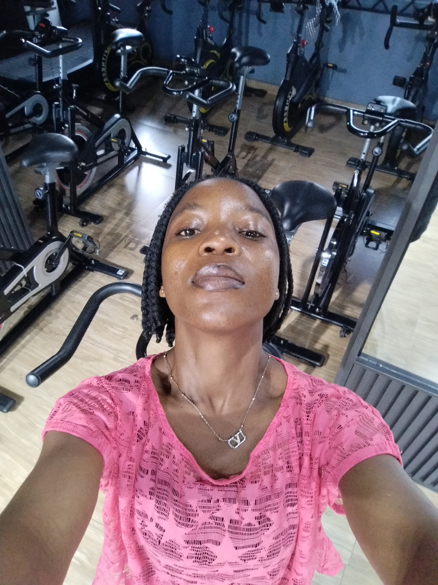 In a gym located in Uselu, Nigeria, a woman of African descent is taking a selfie. She is positioned in the foreground, facing the camera, with a slight upward tilt of her head. She has dark skin, braided hair that is styled in two long plaits falling over her shoulders, and is wearing a bright pink, sheer, lace-like top over a dark-colored undergarment. A silver necklace with a pendant shaped like the letter 'V' is visible around her neck.
The background of the image is filled with stationary exercise bikes, commonly known as spin bikes. These bikes are arranged in rows, with some partially visible in the frame. The gym floor appears to be wooden. To the right of the frame, a reflective surface, likely a mirror or glass panel, shows a distorted reflection of the gym environment and possibly other equipment. The lighting in the gym appears to be artificial, creating some shadows and highlights on the equipment and the floor. The overall environment suggests a fitness facility in operation.
