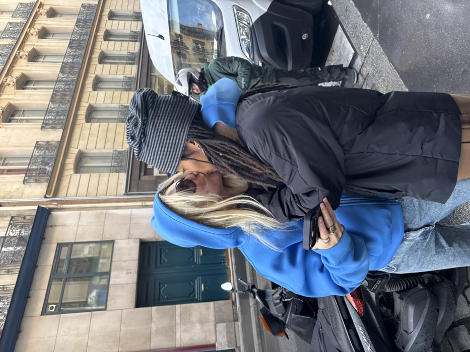 A couple is kissing on a city street. The person on the right has dreadlocks and is wearing a gray and black striped beanie. The person on the left has blonde hair and is wearing sunglasses and a blue hoodie. A white van and a scooter are parked nearby. A building with several windows is in the background. They are in love.