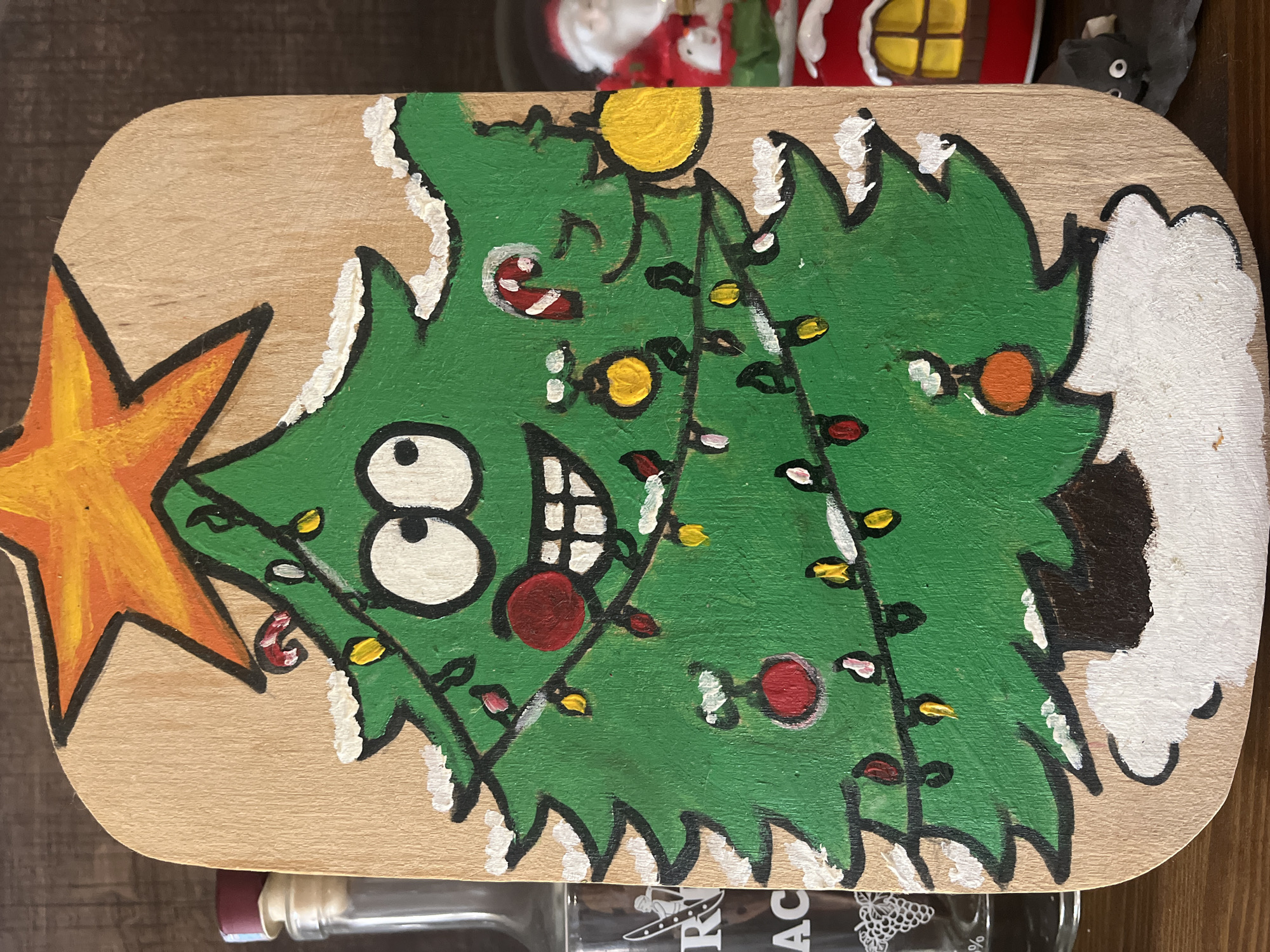 A hand-painted wooden Christmas tree decoration is displayed on a wooden surface. The tree is depicted with a cartoonish face, wide eyes, a large smile with teeth, and rosy cheeks. It is adorned with colorful Christmas lights, ornaments, and candy canes. White paint is used to represent snow on the branches and at the base of the tree. A yellow star with orange accents sits at the top of the tree. To the left of the decoration is a glass bottle with a dark liquid and a cork. To the right, a small figurine of Santa Claus and a red house with a yellow window are visible. The background is dark and out of focus. The location is not specified in the image or surrounding context.