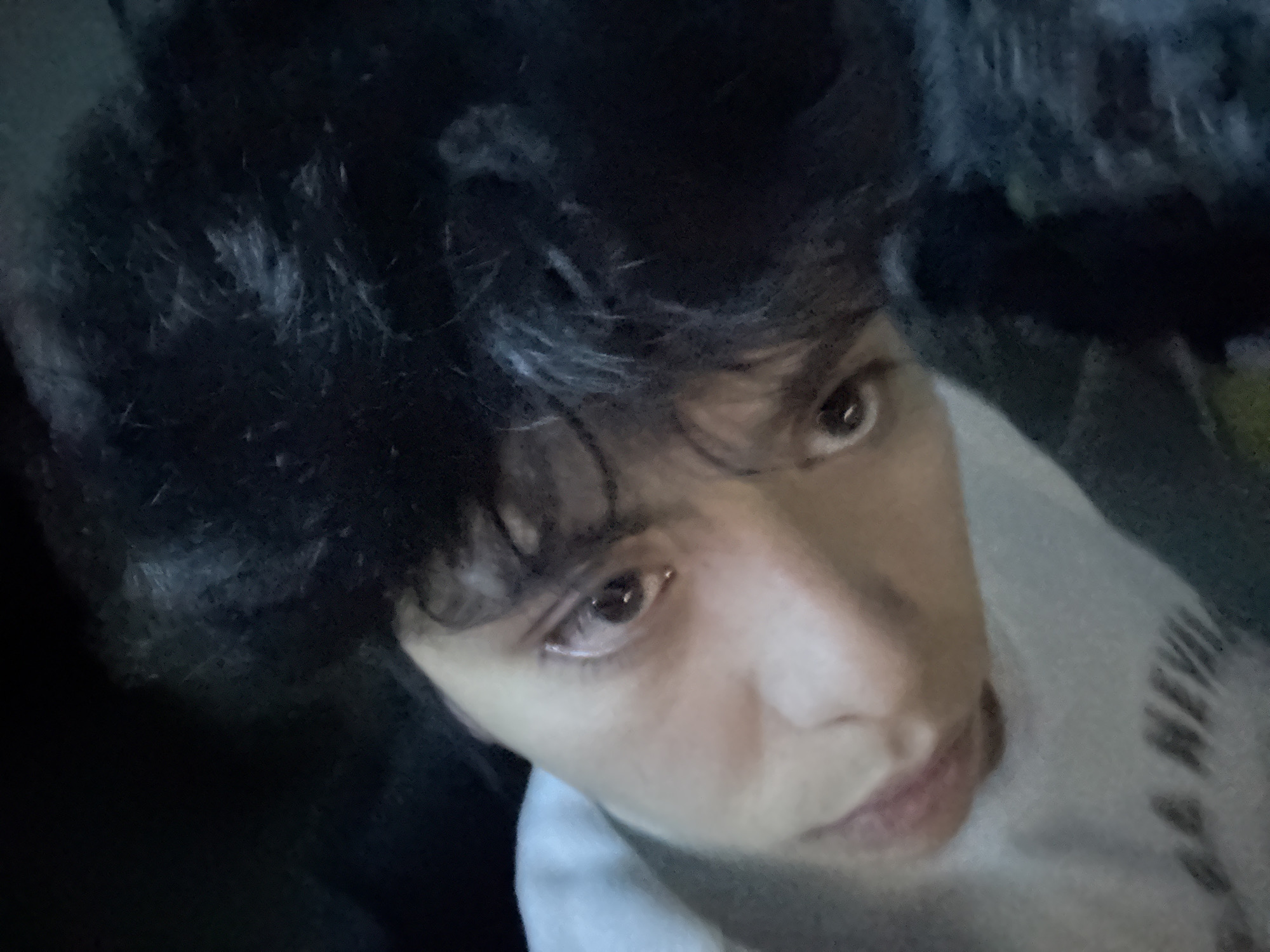 A close-up, angled selfie of a person with dark, curly hair. The person is wearing a white garment with black letters partially visible. The location is unknown.