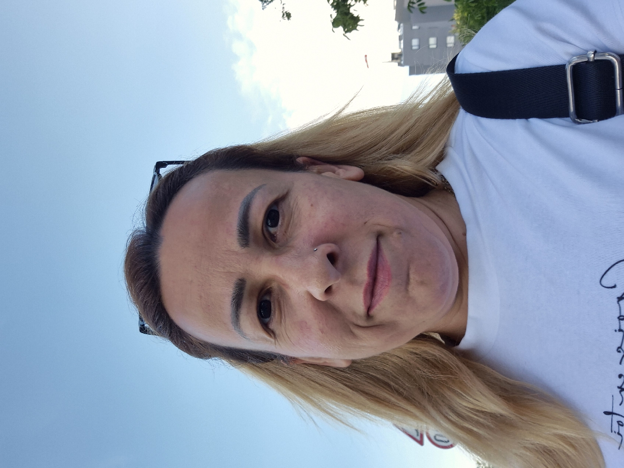 The media file is a close-up, self-portrait of a woman outdoors. She has blonde hair, and is wearing a white t-shirt with a black strap across it. She is looking towards the camera with a slight smile. A small nose ring is visible. The background consists of a bright blue sky with some scattered clouds, and a building and some trees are visible in the upper right corner, suggesting an urban or suburban setting. The lighting suggests it is daytime, possibly late afternoon given the quality of light. The woman appears relaxed and happy. No discernible text is visible in the image.