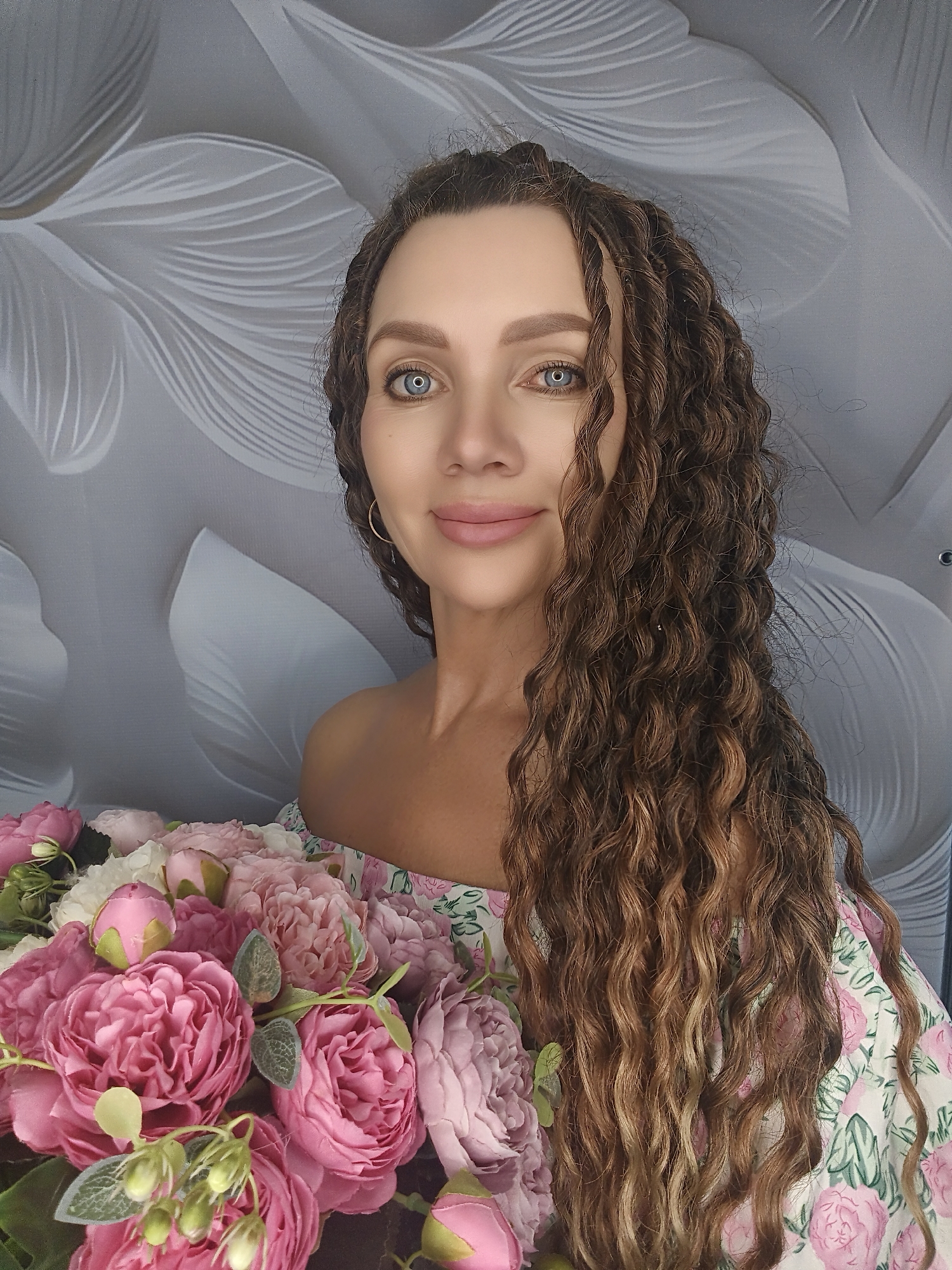 A medium shot shows a woman with long, curly brown hair and blue eyes, smiling at the camera. She is wearing an off-the-shoulder dress with a floral pattern and holding a bouquet of pink and white flowers. The background is a gray wall with a white floral pattern.