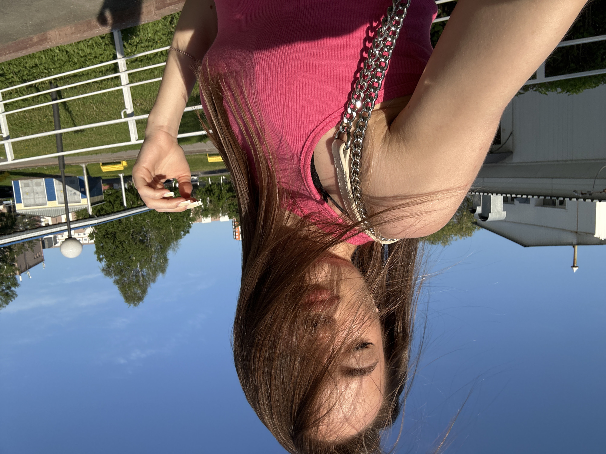 A young woman is depicted in a close-up, head-and-shoulders portrait, taken from a slightly low angle outdoors on a sunny day. She has long, straight dark brown hair, some of which falls across her face, obscuring part of her eyes. Her lips are slightly parted. She is wearing a fitted, ribbed pink tank top. A silver chain strap of a shoulder bag is visible on her right side, resting on her shoulder and chest. Her right arm is raised, and her hand, adorned with long, light-colored artificial nails, holds a small, light-colored object, possibly a vape or similar device. A delicate bracelet is visible on her wrist.
The background shows a clear blue sky with a few faint clouds. Below the sky, on her right, there are white buildings. On her left, a white railing separates the foreground from a lower grassy area. In the distance beyond the grass, there are more buildings, including one with blue and yellow sections, and green trees. A street light is also visible. The bright sunlight creates clear shadows, with one appearing to be cast by a person on the ground near the railing. The overall scene suggests a pleasant outdoor setting, likely during the day, given the strong illumination. There is no visible text in the image.