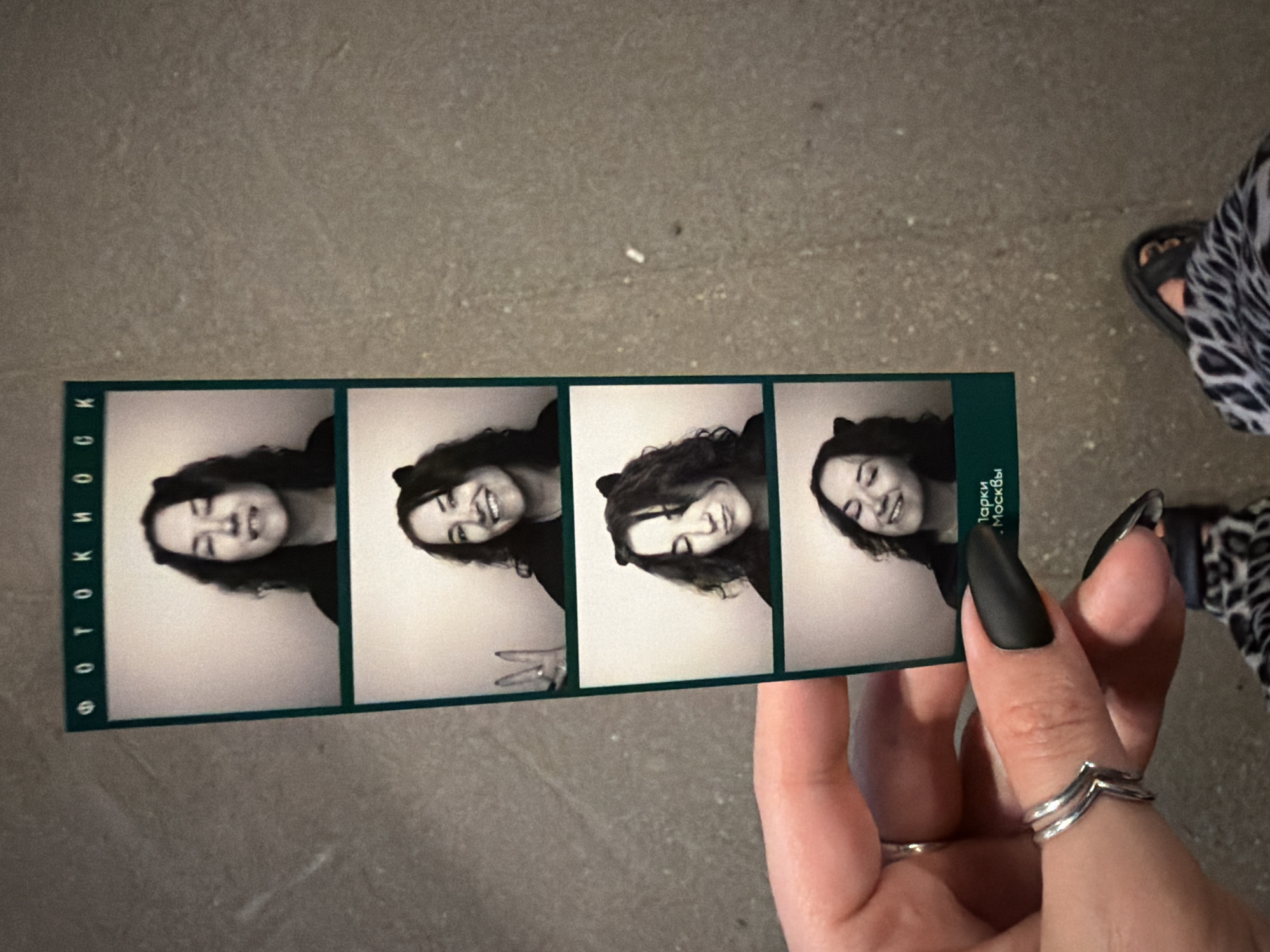 A grayscale photograph depicts a hand holding a photo booth strip featuring four images of the same smiling woman. The hand has long, pointed matte black nails and wears two silver rings. The photo strip has text in Russian. The woman in the photo is also smiling, and in one photo, she is making a peace sign with her fingers. The background behind the hand and photo strip is a gray textured surface. A person is visible in the background, wearing animal-print pants and sandal. The text "Парки Москвы" indicates the location of Unknown, Unknown is Moscow Parks.