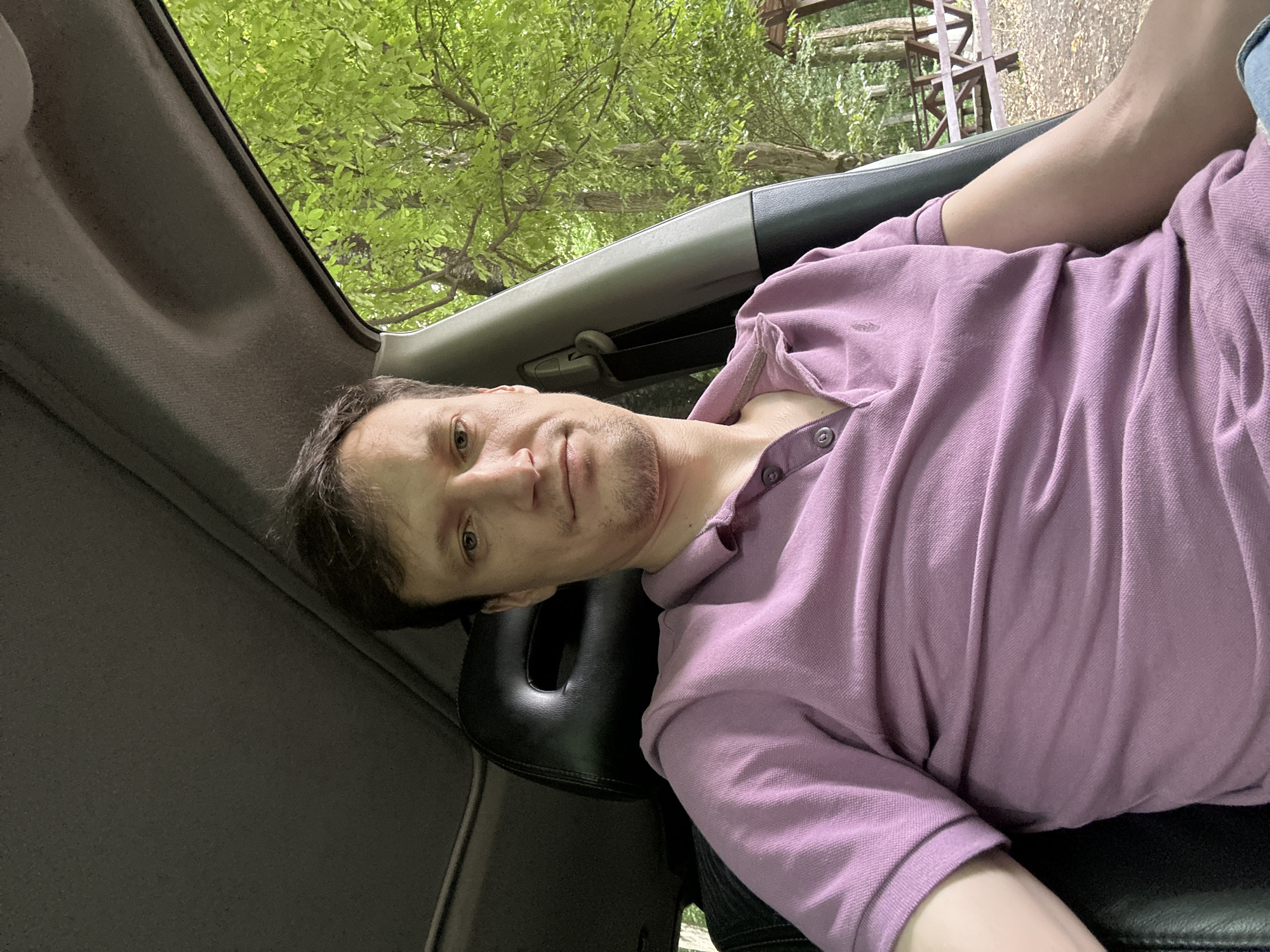 A selfie taken from inside a car shows a man with short brown hair, blue eyes, and light skin. He is wearing a purple polo shirt with a collar and two buttons at the neck. The man is sitting in the driver's seat, with the car's interior visible around him, including a black headrest and the car's roof lining. Through the open car door on the right, a lush green forest with trees and a wooden picnic table can be seen. The background suggests the car is parked in a natural setting, possibly a park or a wooded area, in or near the city of Shatava, Ukraine. The lighting is natural, indicating it's daytime.