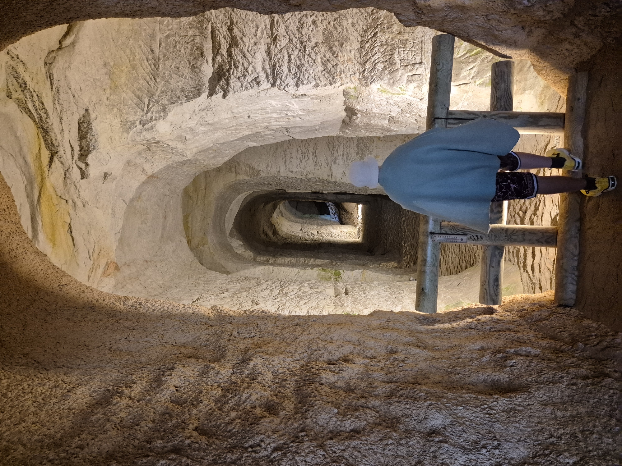 A person wearing a light blue cloak and a white hat walks through a man-made cavern. The cavern walls are rough and textured, with rough-hewn arches creating a tunnel-like passage that recedes into the distance. Wooden railings line the path. The scene appears to be inside the Piusa Sand Caves in Piusa, Estonia, a network of underground tunnels excavated for glass sand.