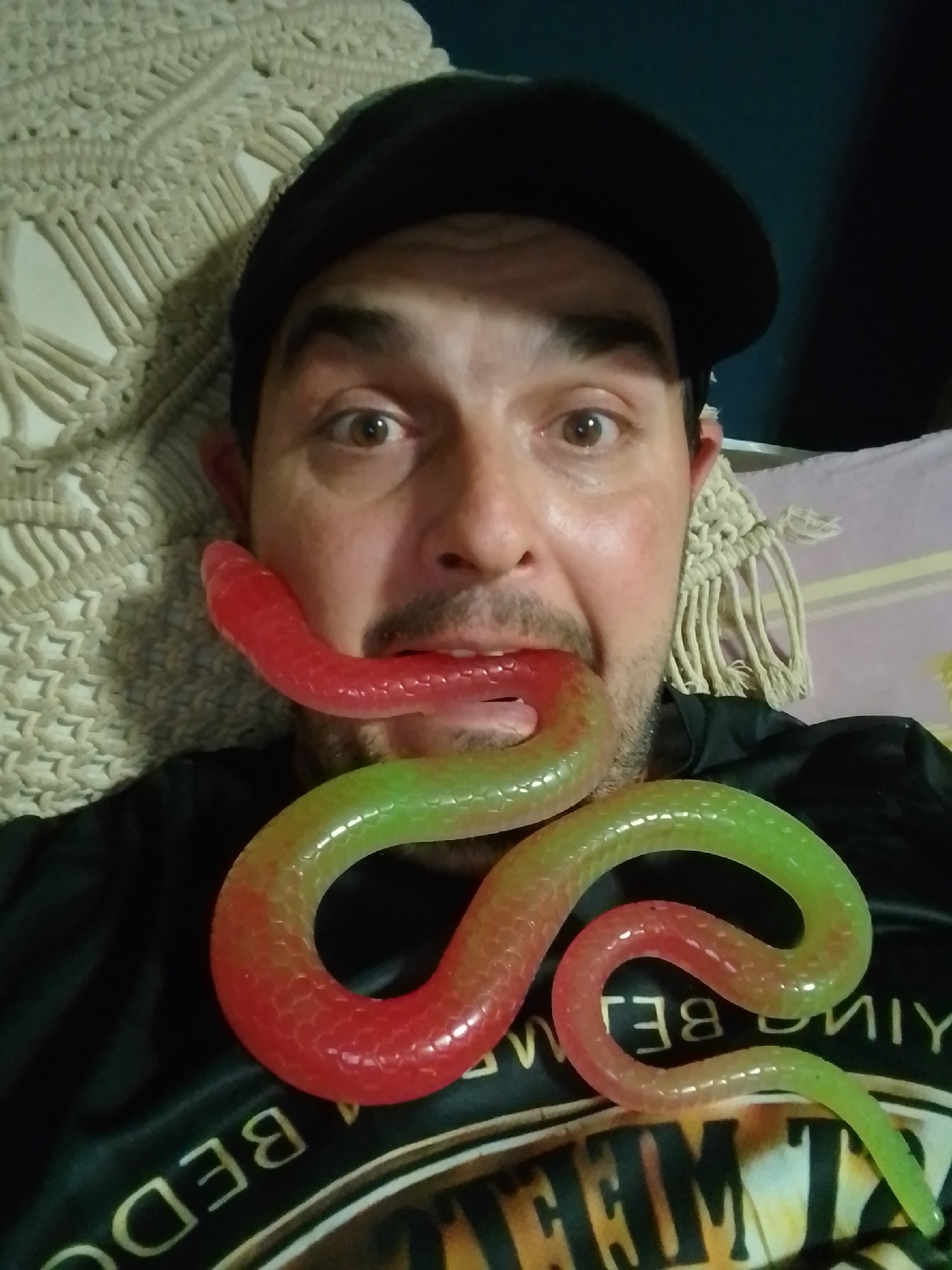 A man wearing a black baseball cap has a red and green gummy snake across his face, with the red end in his mouth. He is lying down on a macrame-style textile, and a pink patterned textile is visible behind him. The man is wearing a black t-shirt with text and a graphic design. This image was captured in the city of Wedel, Germany.