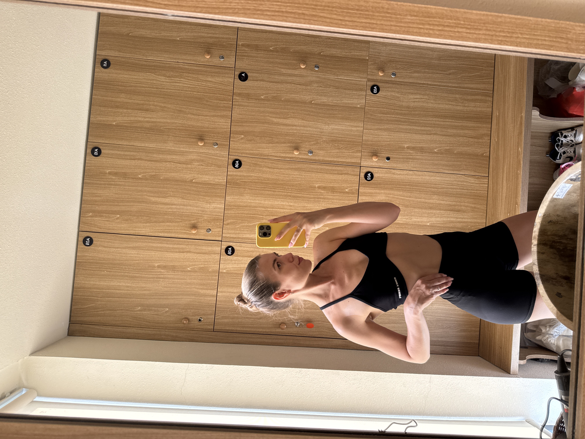 A mirror reflects a fair-skinned woman with blonde hair pulled into a bun, wearing a black sports bra and black shorts, holding a yellow phone. She is positioned in front of a wall of wooden lockers with black numbered labels. A white window is located below the lockers. The setting is a changing room in Unknown, Unknown.