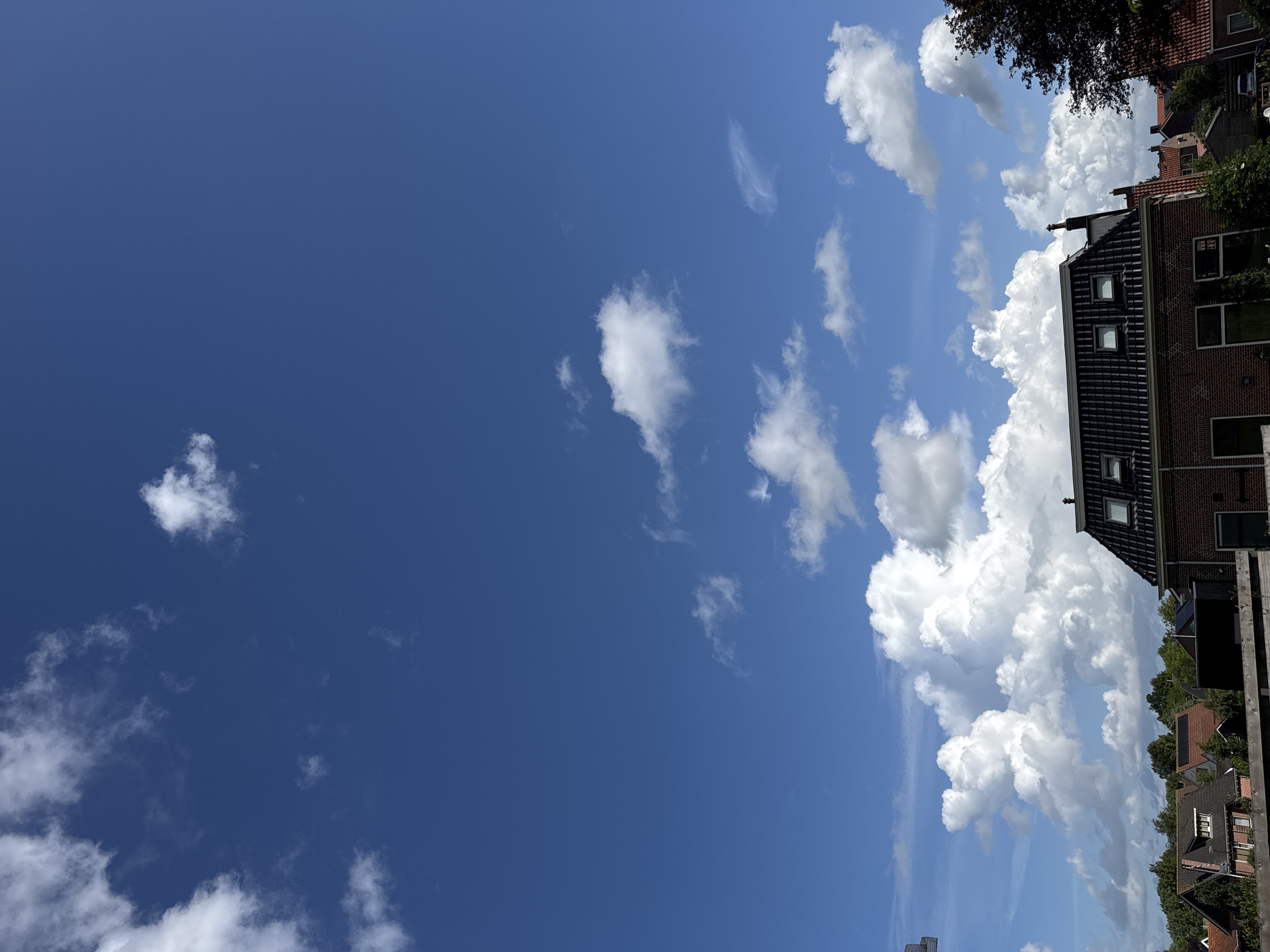 The image shows a suburban street scene under a bright blue sky with scattered white cumulus clouds. In the foreground, a dark-tiled roof of a house is visible, featuring several dormer windows. The house is constructed of red brick. To the left and behind this house, other residential buildings with various rooflines and colors can be seen, interspersed with lush green trees. The overall impression is a peaceful, sunny day in a residential area.