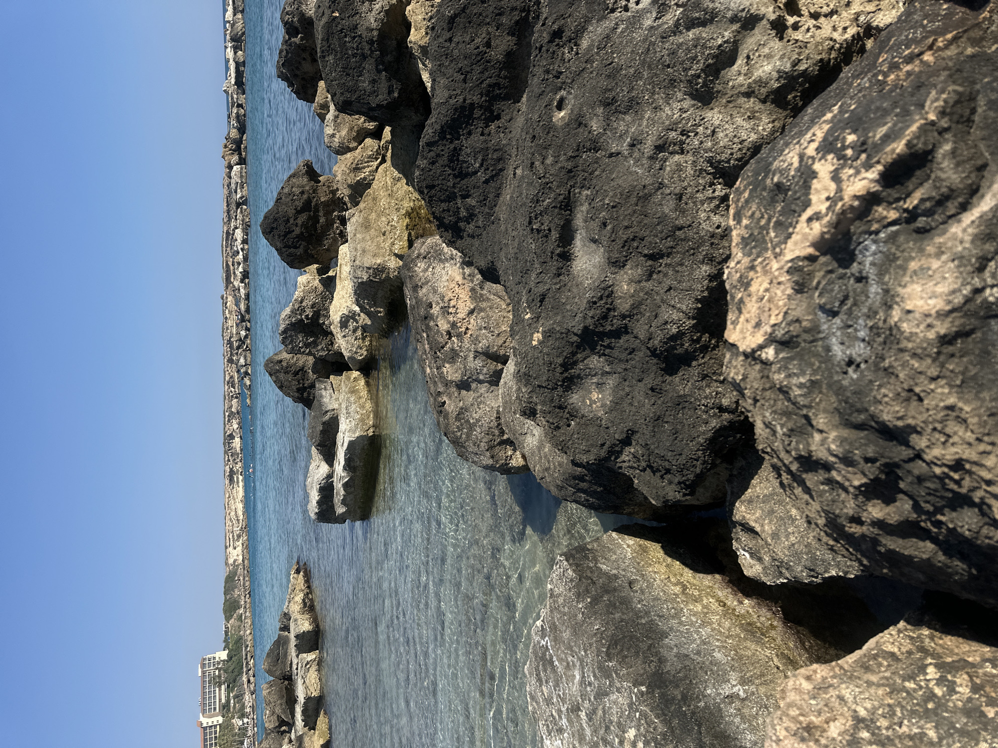 A low-angle, wide shot captures a sunny seascape in Peyia, Cyprus. In the foreground, large, dark volcanic rocks dominate the frame, with a few lighter-colored rocks interspersed. The clear turquoise water of the Mediterranean Sea gently laps around the rocks. In the mid-ground, the water stretches towards a distant breakwater constructed of similar rocks. A multi-story, light-colored hotel building is visible on the left side of the horizon, situated on a rocky hillside dotted with sparse vegetation. A few people can be seen in the water near the breakwater, appearing as small dark figures. The sky is a clear, vibrant blue. The overall impression is one of a tranquil coastal scene on a bright day.