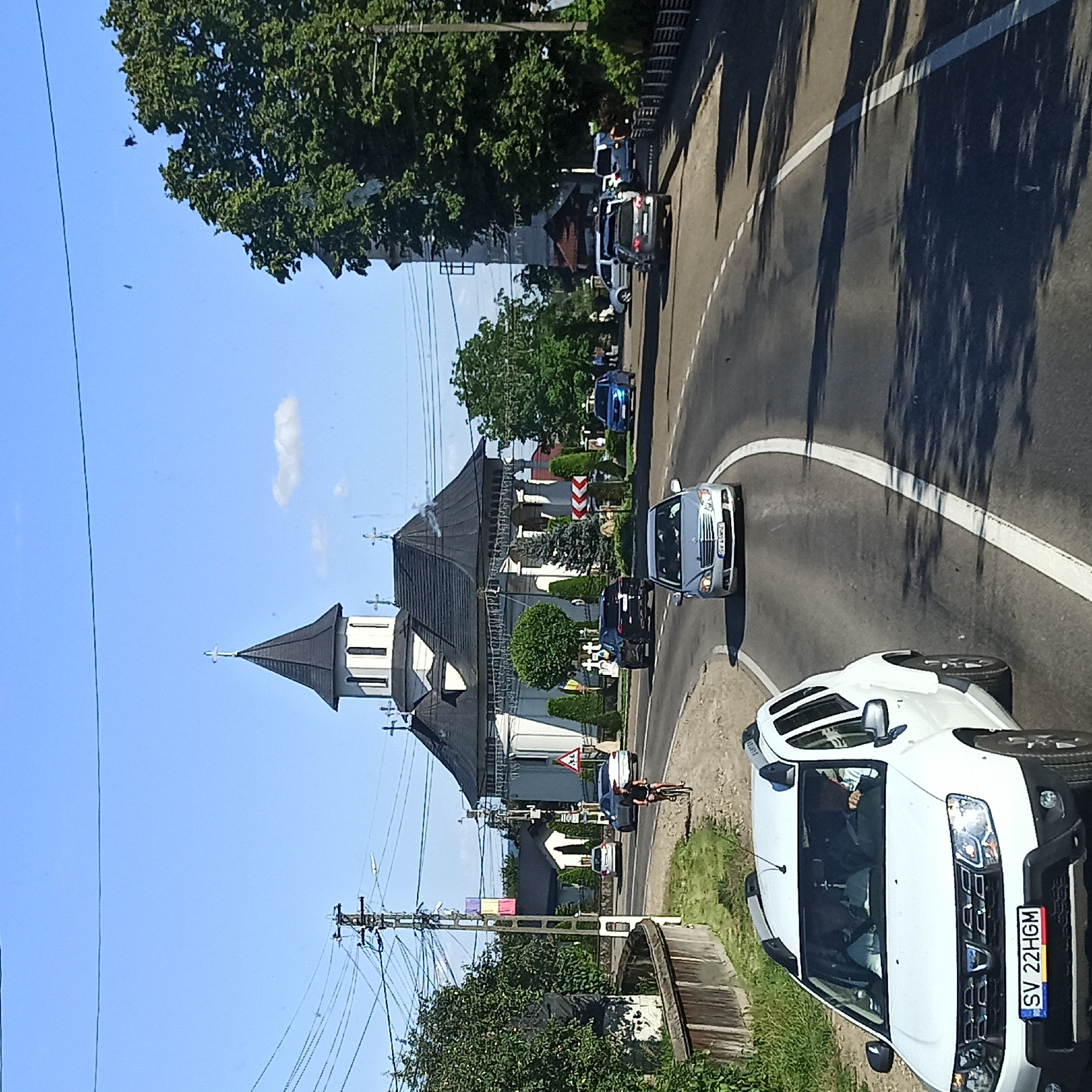 The image captures a sunlit street scene in Milișăuți, Romania, featuring a prominent white church with a dark, multi-tiered roof and a steeple topped with a cross. The church is flanked by trees and smaller buildings. A two-lane asphalt road curves through the scene, with a white line delineating the lanes. Several vehicles are visible: a white Dacia Duster SUV in the foreground with Romanian license plates "SV 22HGM," a silver Mercedes-Benz sedan behind it, a dark SUV, and a blue car further down the road. A cyclist, a person wearing dark clothing, is riding a bicycle on the right side of the road. Power lines and poles are a significant feature of the environment, particularly on the left side of the image. A wooden fence runs along the left edge of the road. The sky is clear blue with a few scattered clouds.