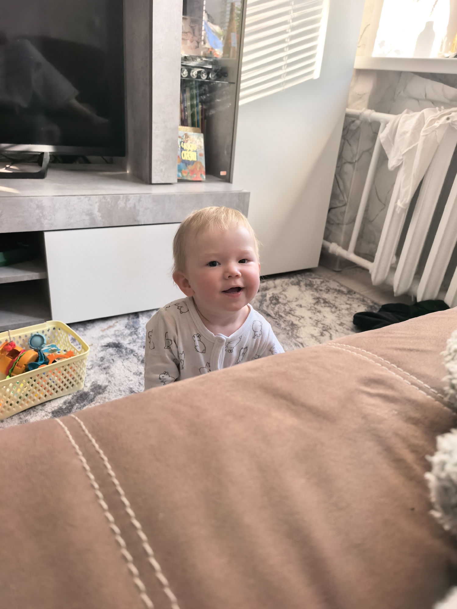 A fair-skinned baby with blonde hair, wearing a white patterned onesie, sits on a rug. A tan couch partially obscures the lower portion of the view. To the left, a yellow basket holds colorful toys. Behind the baby is a light gray carpet, a light gray and white TV stand with a TV, a glass cabinet, and a white wall with a white radiator. A window with blinds is visible in the background.
This scene is likely inside a home, potentially in the city of Sterlitamak, Russia.