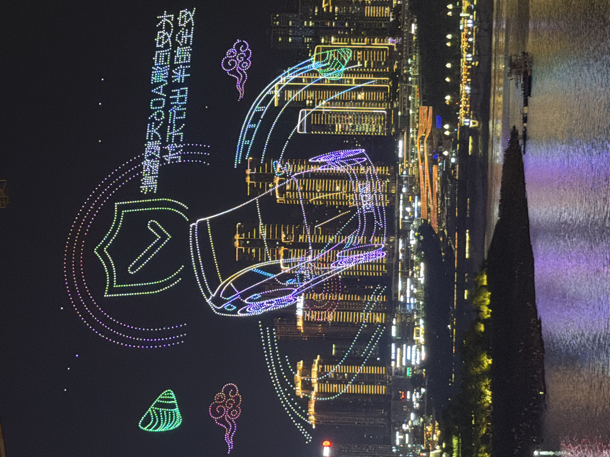 This media file captures a nighttime drone show in Yuzhong District, China. The scene is set against a backdrop of a city skyline illuminated by numerous lights, with a body of water reflecting the colorful display. The drone show itself forms intricate patterns in the sky. One prominent formation is a shield-like shape with an arrow pointing upwards, rendered in green and pink lights. Another significant element is a large, stylized image resembling a traditional Chinese opera mask or a face with exaggerated features, outlined in purple and white lights. Smaller, stylized depictions of a zongzi (a type of glutinous rice dumpling) and a cloud or swirl are also visible. These formations are made up of many small, colored lights, creating a vibrant and dynamic spectacle against the dark sky. The city lights, extending into the distance and across the water, suggest a bustling urban environment. The reflection on the water adds depth and enhances the colorfulness of the drone formations. The overall impression is one of a festive and technologically advanced public event. Text is visible in the upper left quadrant, appearing to be Chinese characters, suggesting a specific message or theme for the drone show. The text reads: "不忘初心 牢记使命 永远跟党走" which translates to "Never forget the original intention, keep the mission in mind, and always follow the party."