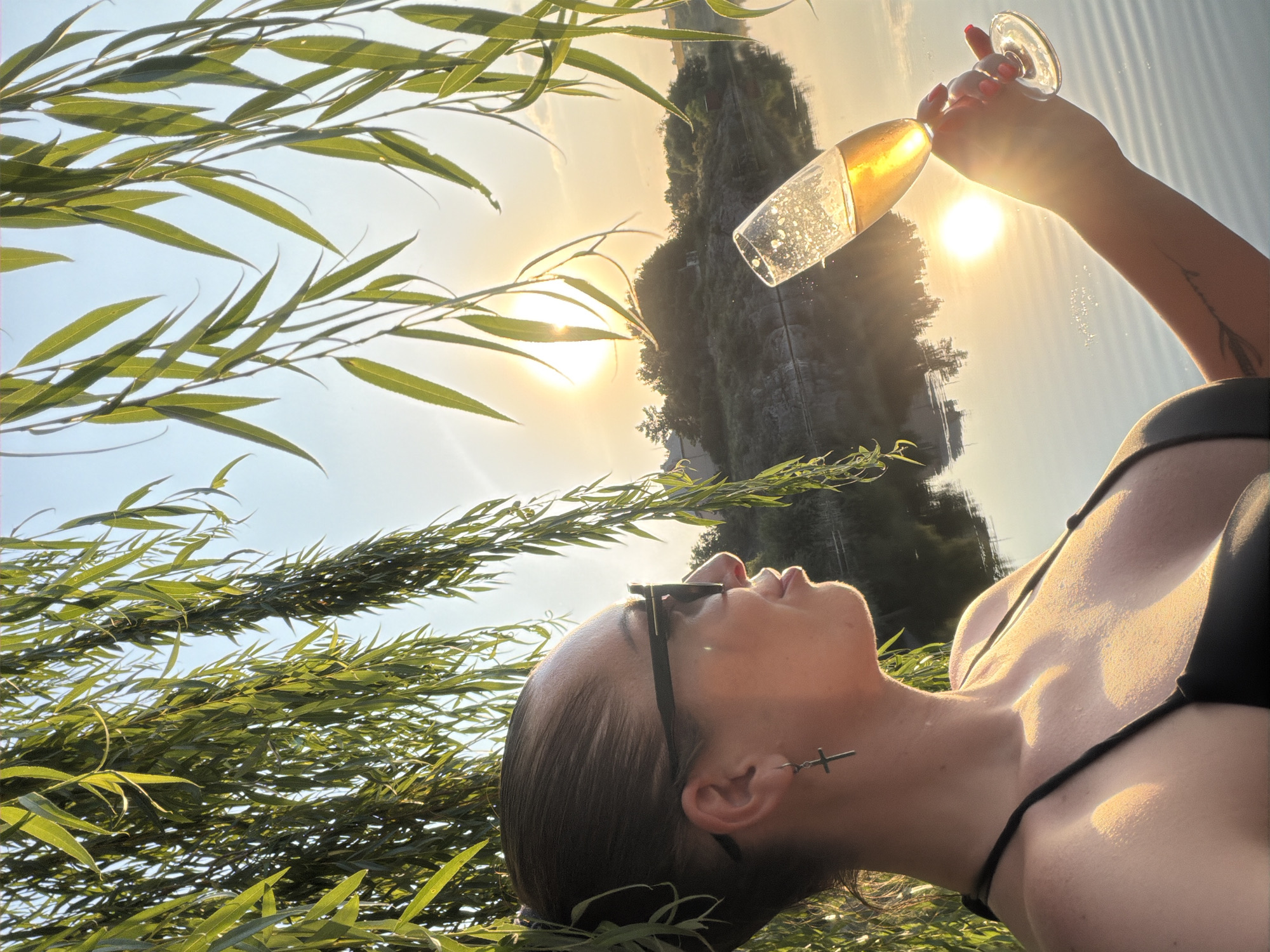 A medium shot shows a woman in a black bikini holding a champagne flute near a body of water, with willow branches framing the scene. The sun is setting behind a hilly, wooded area across the water, casting a golden glow. The woman is looking to her left, her face illuminated by the sunset. She is wearing sunglasses and a silver cross earring. The champagne flute is tilted, with bubbles visible in the golden liquid. The water reflects the sky and the surrounding landscape. The location is identified as Lozuvatka, Ukraine.