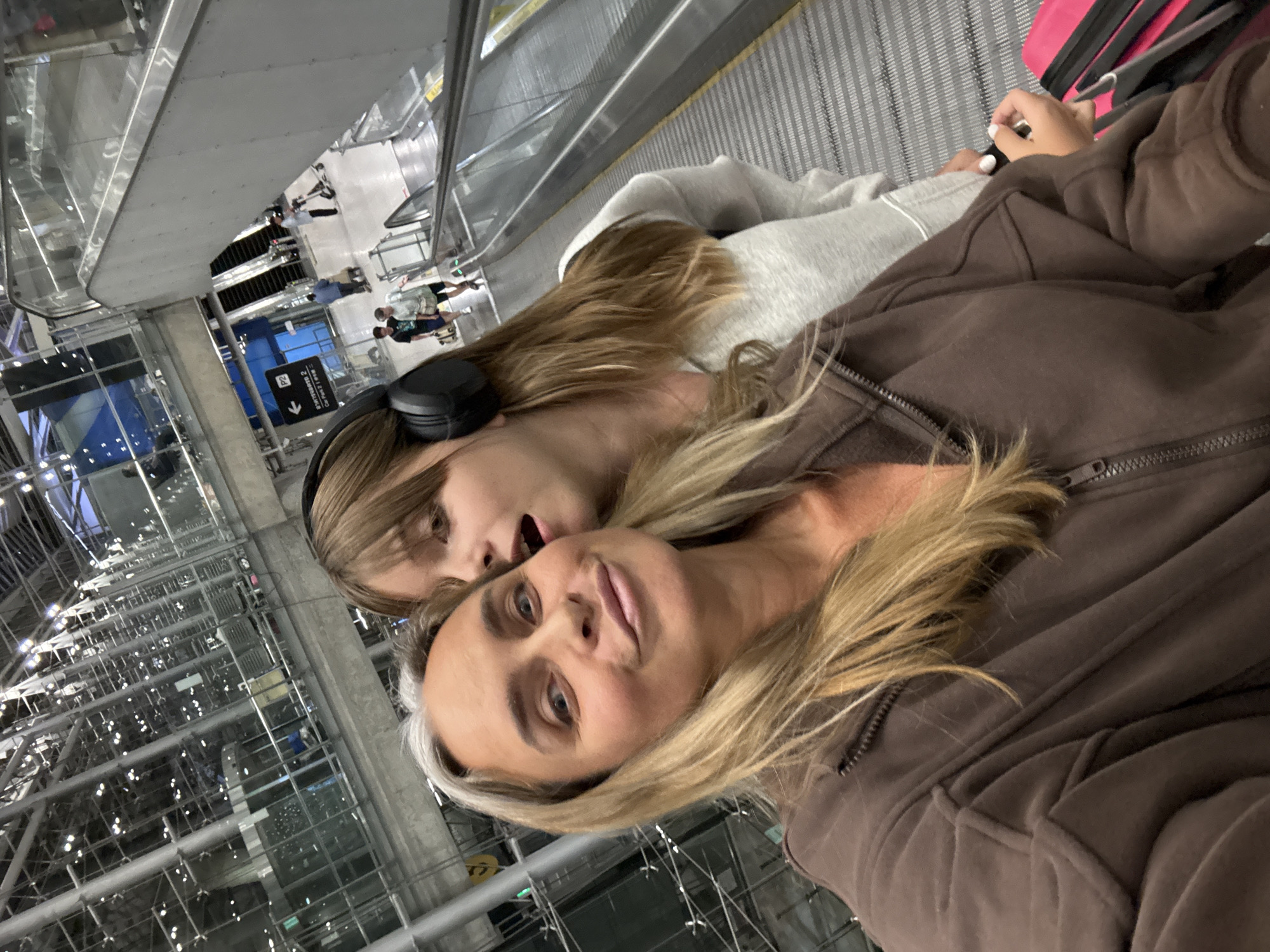 This is a selfie taken by two females on an escalator in an airport. The woman in the foreground has blonde hair with gray streaks and is wearing a brown hooded jacket. The woman behind her, who appears to be a teenager, has long brown hair, is wearing headphones, and is leaning in to whisper something to the woman in front. The background shows the interior of a large, modern airport with escalators and other travelers. The image is likely taken in Suvarnabhumi Airport, which is located in the Nong Prue Subdistrict of Bang Phli District, Samut Prakan Province, Thailand.