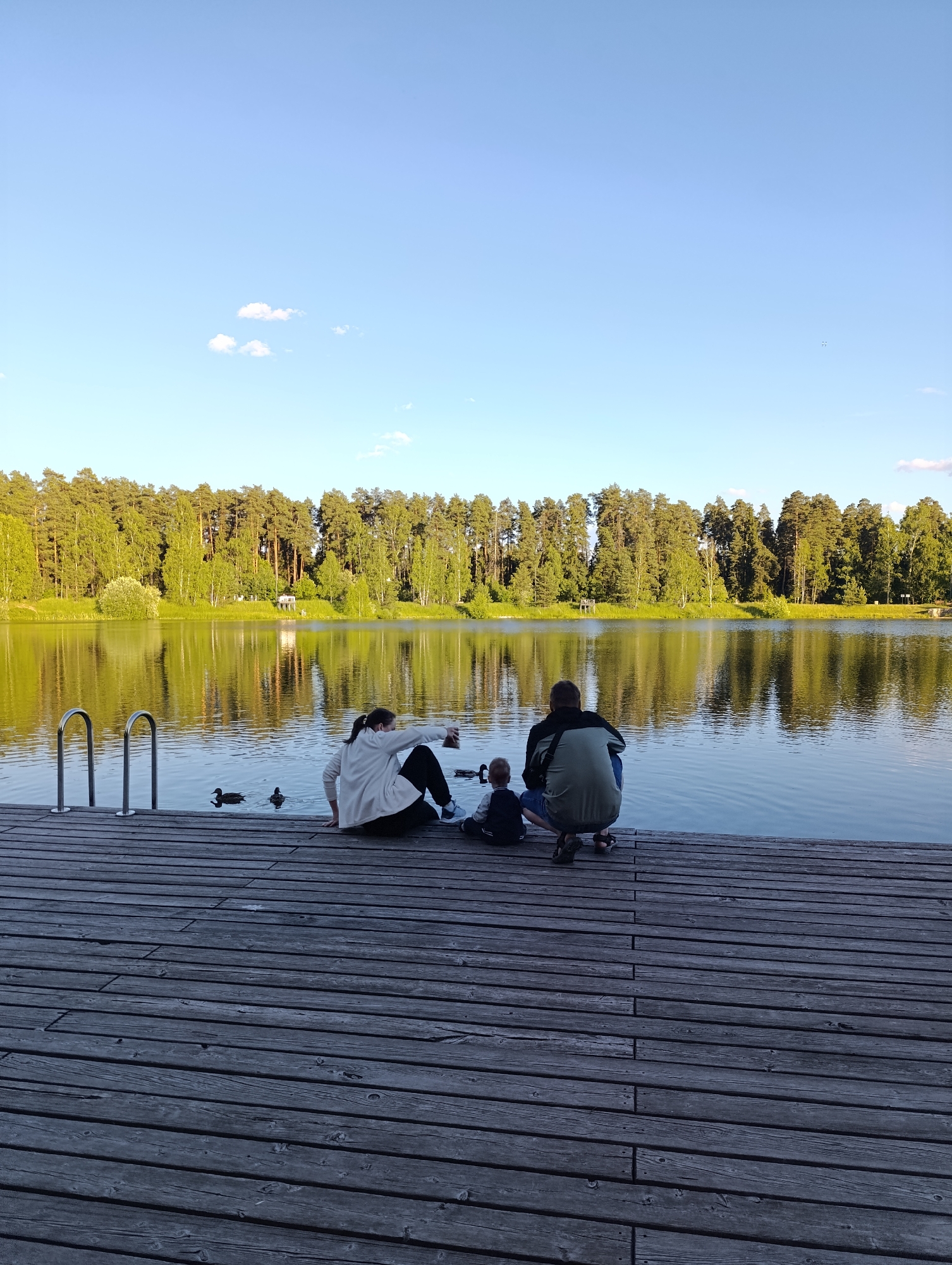 This image depicts a serene scene on a wooden pier by a lake, likely in Chernogolovka, Russia. Three people, two adults and a child, are seated on the pier, facing the water and a dense forest that lines the opposite shore.
The adult on the left is a woman wearing a white hoodie and black pants, facing away from the camera and looking towards the water. Next to her, a small child in a dark jacket and pants is also seated, looking out at the lake. On the right, another adult, presumably a man, is crouched down, also facing the water. He is wearing a green and black jacket and dark shorts. Their postures suggest a relaxed and contemplative mood.
Several ducks are visible swimming in the calm water near the pier, adding to the peaceful atmosphere. The lake reflects the clear blue sky and the lush green trees of the forest, creating a picturesque backdrop. A few small, fluffy clouds dot the sky. The sun appears to be low in the sky, casting a warm, golden light, suggesting it is either early morning or late afternoon. The overall impression is one of tranquility and family enjoyment of nature. No text is visible in the image.