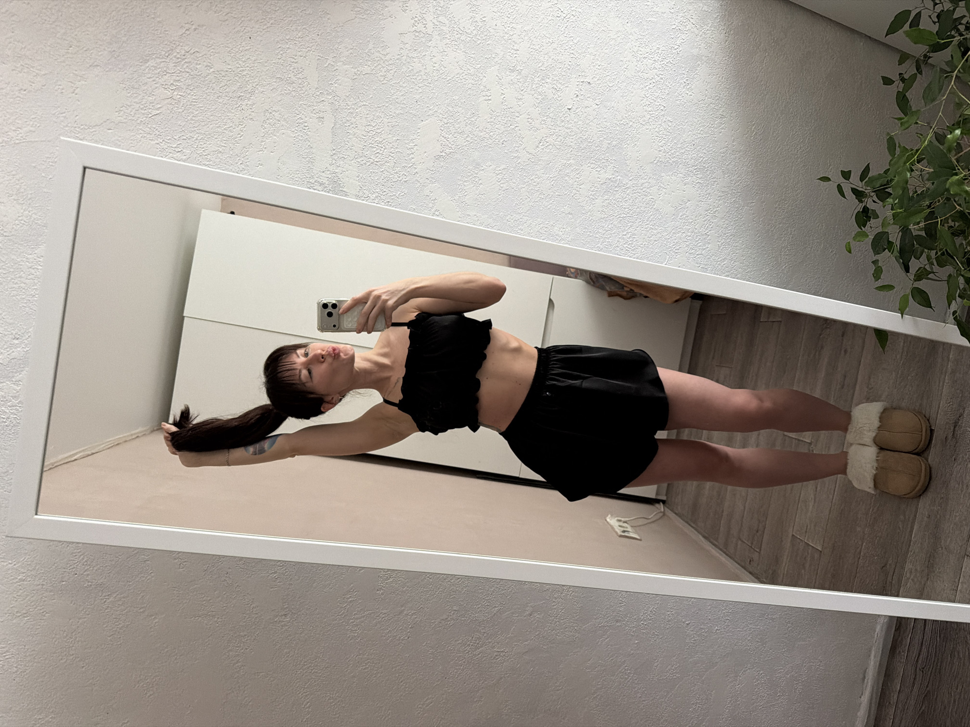 A young adult female is taking a selfie in a mirror. She is dressed in a black two-piece outfit consisting of a top and shorts. She is wearing beige slippers. The background includes a white wall with a textured surface, a white cabinet, a plant, and a wooden floor. The image appears to be taken indoors, possibly in a home in Perm, Russia.