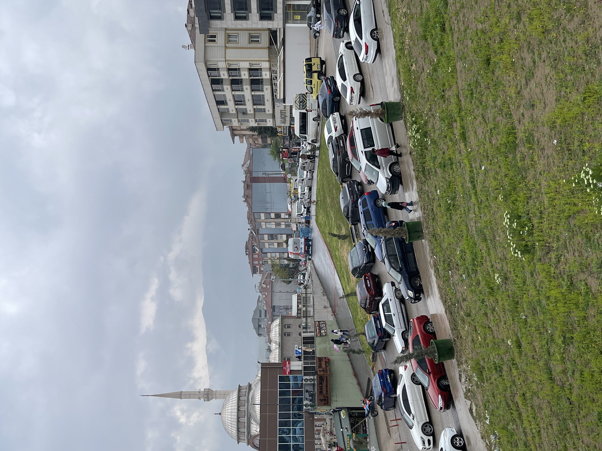 The image depicts a street in Karabük, Turkey, with a mosque visible in the foreground. The street is lined with parked cars of various colors and models, interspersed with green planter boxes. A grassy area runs alongside the road, dotted with white flowers. Several buildings are visible in the background, ranging from low-rise commercial structures to taller apartment blocks. The sky is overcast with a gray hue. Pedestrians are seen walking along the street. The architecture and landscaping suggest an urban or suburban setting.
