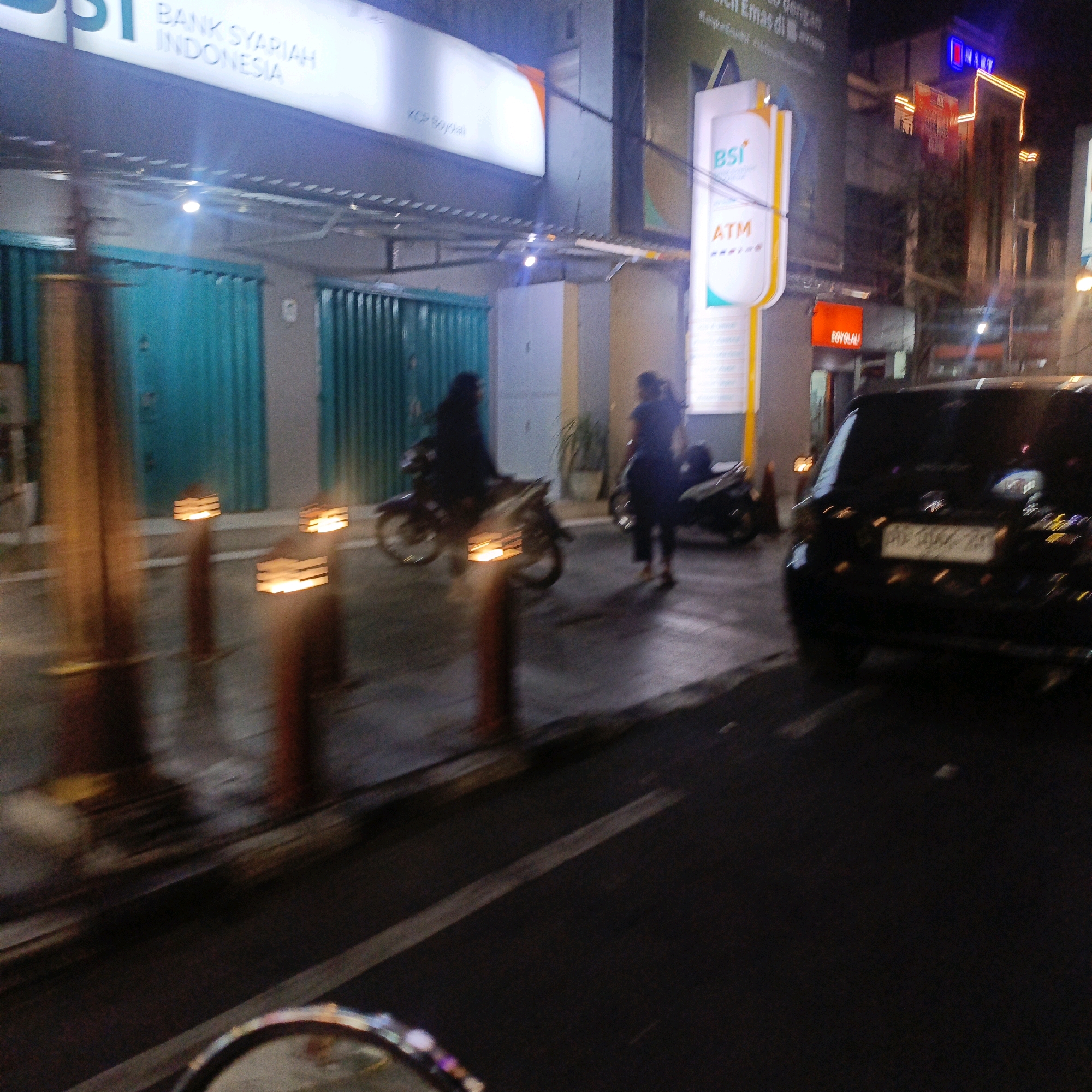 The image depicts a street scene at night in Boyolali, Indonesia. In the foreground, a black car is visible from the rear, its license plate partially obscured. To the left, a motorcycle with a rider is parked. Further down the street, another motorcycle is parked, and a person is standing next to it. The background features a building with a prominent sign that reads "BANK SYARIAH INDONESIA," and another sign for "BSI ATM." Several lamps with a warm glow are positioned along the sidewalk. The overall atmosphere is that of a typical urban environment at night.