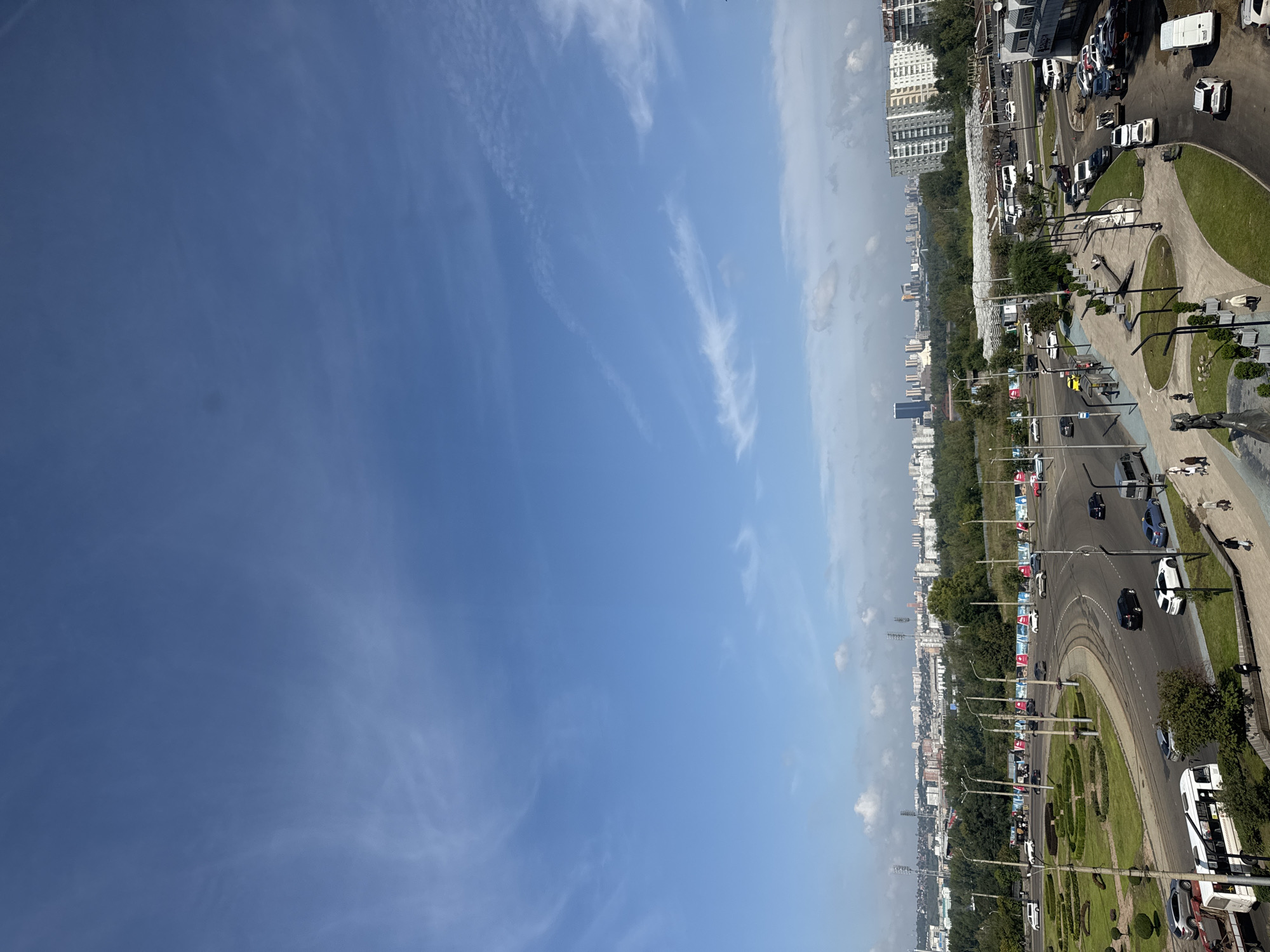 The image captures a wide shot of a cityscape in Krasnoyarsk, Russia, dominated by a vast blue sky with wispy white clouds. In the foreground, a busy intersection features a multi-lane road with various vehicles, including cars, a bus, and what appears to be a trolleybus. Greenery is abundant, with manicured circular garden beds and trees lining the roads and sidewalks. Pedestrians are visible on the sidewalks, some walking and others standing. In the mid-ground, more trees and buildings form a dense urban landscape, interspersed with what looks like a greenhouse structure. The background reveals a sprawling city with numerous multi-story buildings and infrastructure, including what might be industrial structures or stadiums in the distance. The overall impression is a sunny day in a developed urban area.