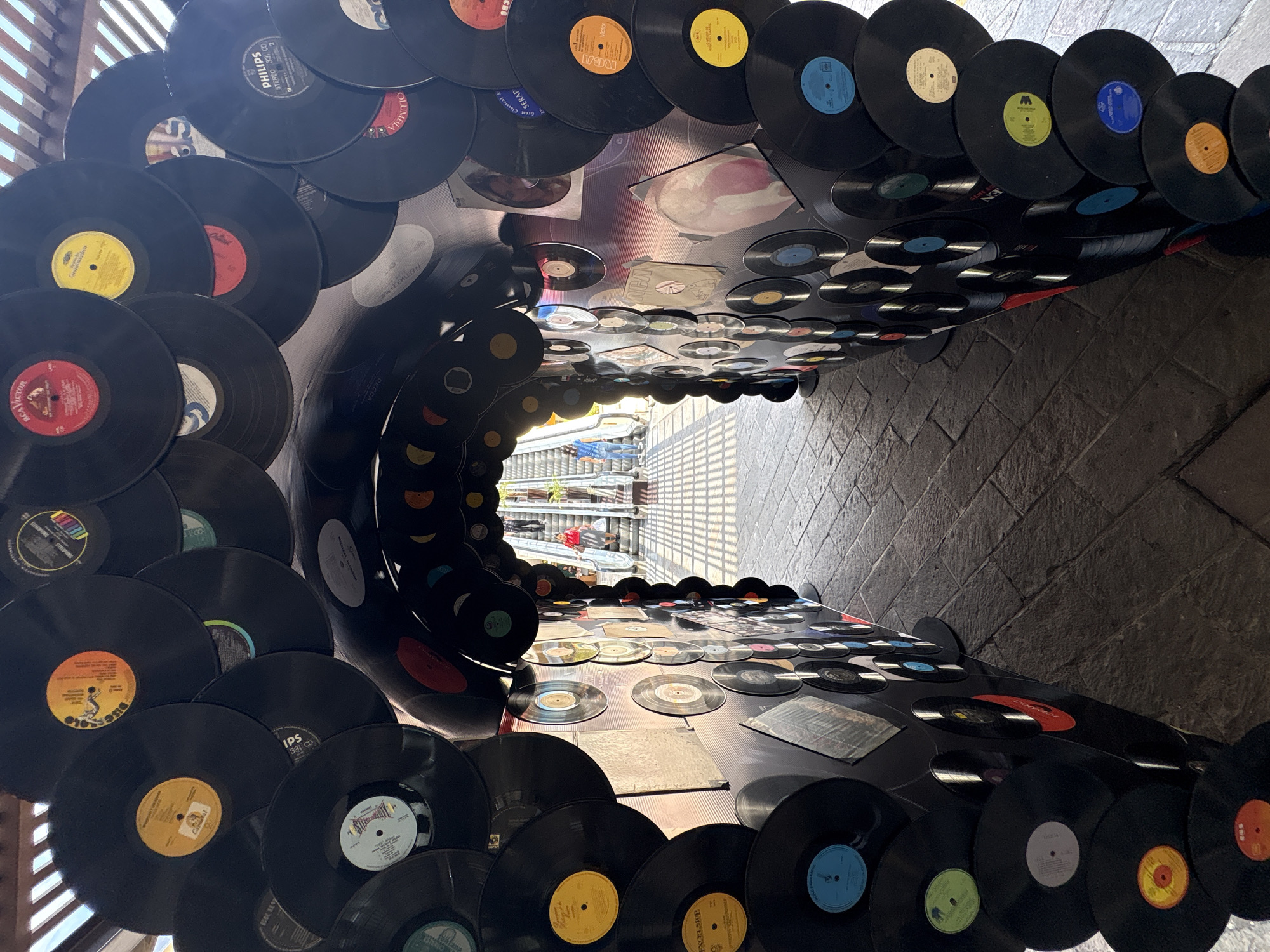 The image shows an art installation made of vinyl records, forming a tunnel-like structure. The records are black and have various colored labels, some of which are visible and legible, displaying text like "RCA Victor," "Philips," and "Columbia." The tunnel is illuminated by natural light filtering through an overhead structure that appears to be made of wooden slats. Inside the tunnel, an escalator is visible with people ascending. One person in the foreground of the escalator is wearing a red top and a striped skirt, carrying shopping bags. Another person further up the escalator is wearing a blue top and jeans. The ground around the installation is paved with stone blocks. The installation is located in Cuernavaca, Mexico, specifically at the Mercado de Artesanías, where it serves as an entrance or pathway decorated with vintage vinyl records. The overall impression is a creative and retro-themed display, blending art, music history, and a bustling urban environment.