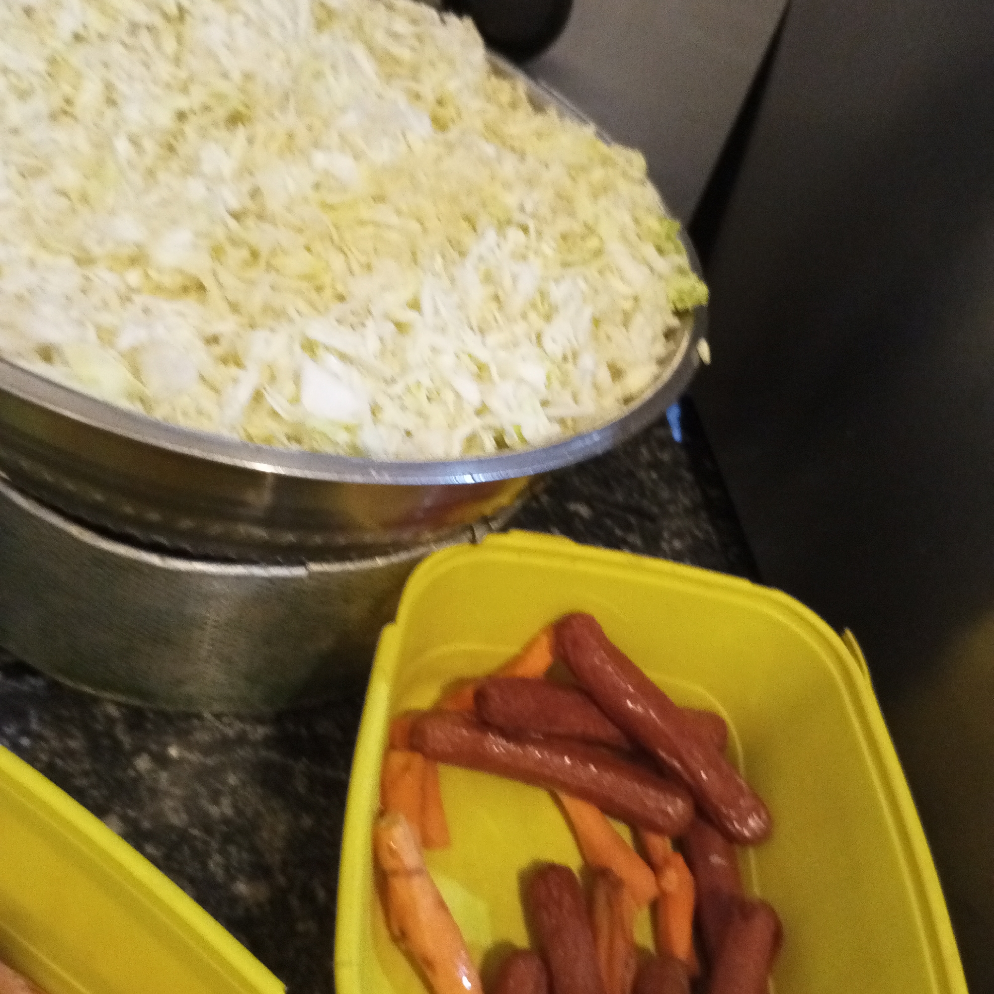 The image captures a food preparation scene in Howayi, Nigeria. A large, reflective metal steaming pot or bowl occupies the upper portion, brimming with a substantial quantity of finely shredded, pale green and white cabbage or similar vegetable. Below and to the right, a vibrant yellow plastic rectangular container holds an assortment of dark reddish-brown, cooked hot dogs or sausages, intermingled with several bright orange, tapered carrot pieces. The objects rest on a dark, speckled countertop surface.