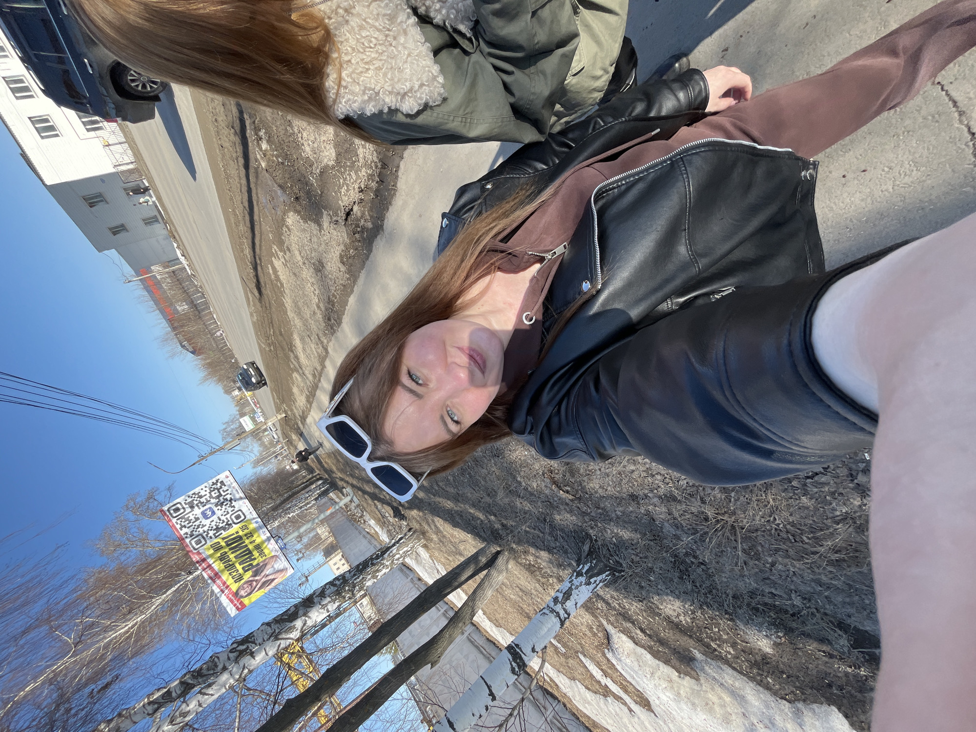 A medium-shot, eye-level outdoor photograph shows two young women taking a selfie while standing next to a sidewalk in Chaikovsky, Russia.
One woman, closer to the camera, wears a brown hooded sweatshirt and a black leather jacket. She has sunglasses with white frames on her head. The other woman, partially visible, wears a green jacket with a white fur-lined collar.
The sidewalk is adjacent to a street with sparse patches of melting snow and vegetation. Across the street, there's a building and bare trees. A banner with a QR code and advertisement for "Radio 7" is attached to a crane and trees. Power lines stretch across a clear blue sky. A car is seen driving down the road.