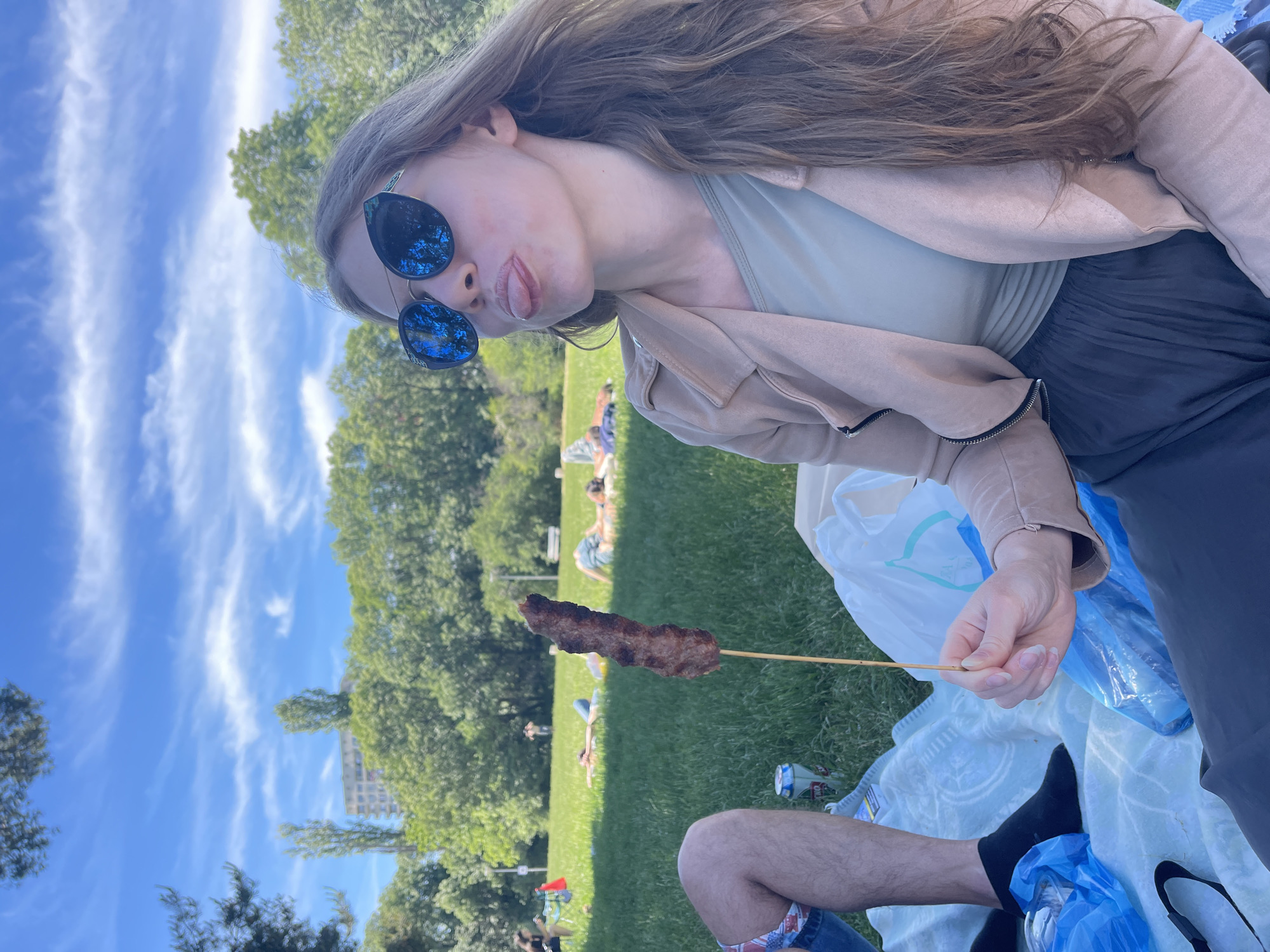 A young woman with long brown hair and sunglasses is pictured holding a shish kebab on a stick. She is wearing a tan jacket over a light-colored top and dark pants. The scene appears to be a picnic on a grassy area with people sunbathing in the background. A blue sky with wispy clouds is visible. The location is likely in a park, given the grassy expanse and trees. The picture was taken in the city of Sofia, Bulgaria.