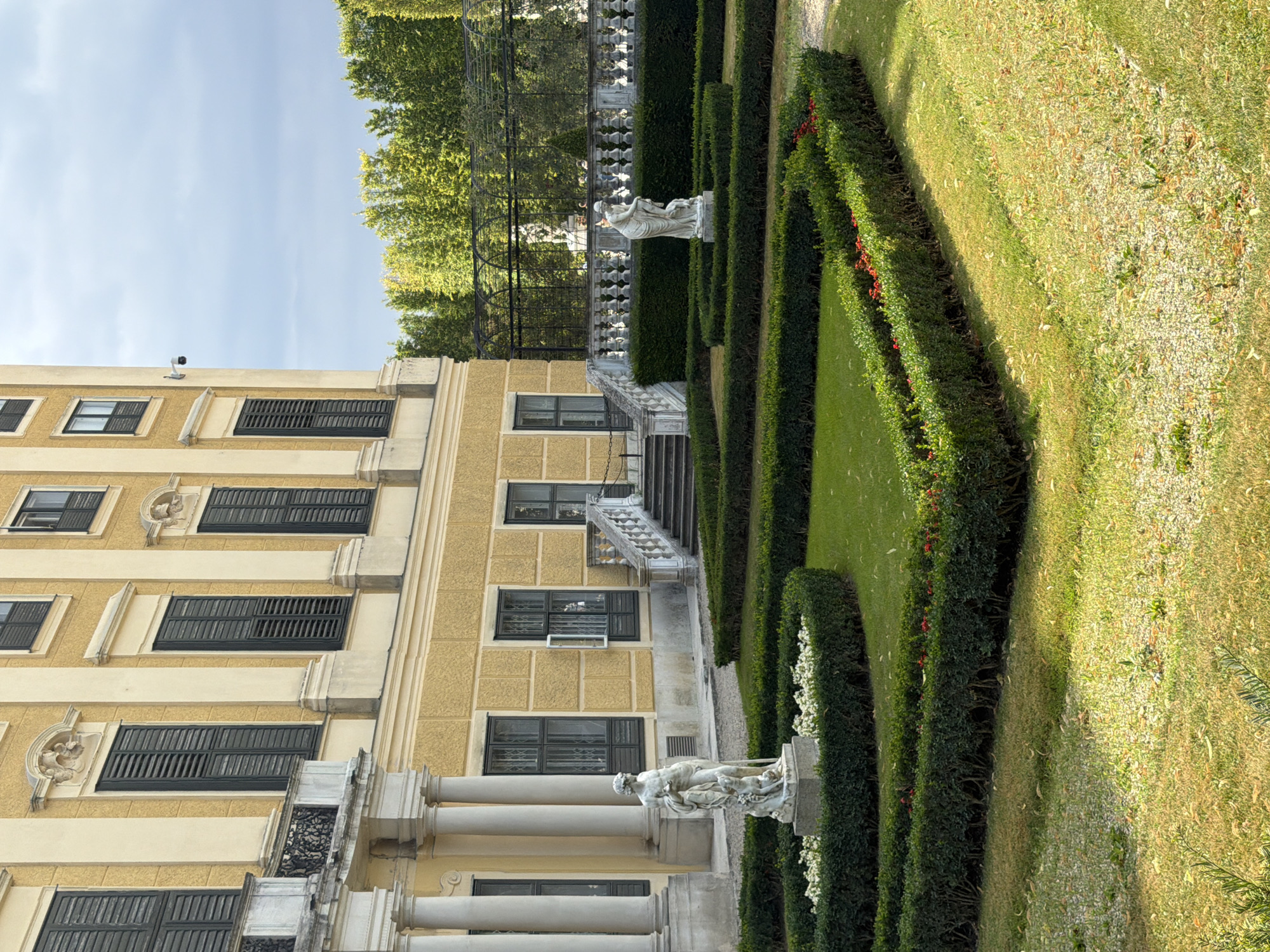 The image depicts a section of the gardens at Schönbrunn Palace in Vienna, Austria. In the foreground, a meticulously maintained formal garden features geometric hedges, lush green grass, and beds of white and red flowers. Two classical white marble statues are prominent within the garden. The statue on the left, positioned in front of a curved hedge, depicts a male figure with a staff and a dog. The statue on the right, situated in the middle ground, shows a female figure with a child.
Behind the garden, a grand, yellow building with large, dark-shuttered windows and classical architectural elements rises. A wide, stone staircase with a decorative balustrade leads up to an entrance on the building's facade. A security camera is visible mounted on the building wall. Beyond the building, a dense line of green trees forms a backdrop, and a black metal fence with ornate railings runs horizontally across the middle ground, separating the palace gardens from the treeline. The sky above is partly cloudy. The overall scene conveys a sense of historical grandeur and formal landscaping characteristic of Viennese palaces.