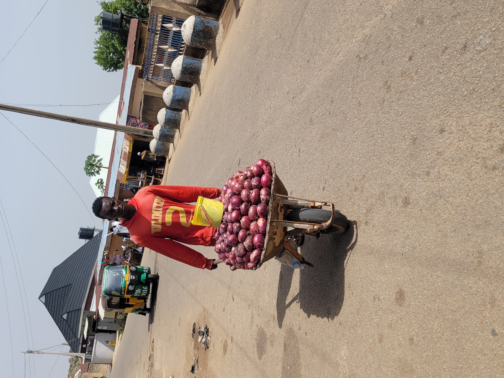 A man pushes a wheelbarrow filled with red onions down a street in Jos, Nigeria. The man is wearing a red long-sleeved shirt with yellow lettering. A yellow container sits among the onions. The wheelbarrow is casting a shadow on the pavement. The street is paved and lined with concrete bollards. Buildings are visible in the background, including a green and yellow motorized tricycle. Power lines are overhead.
