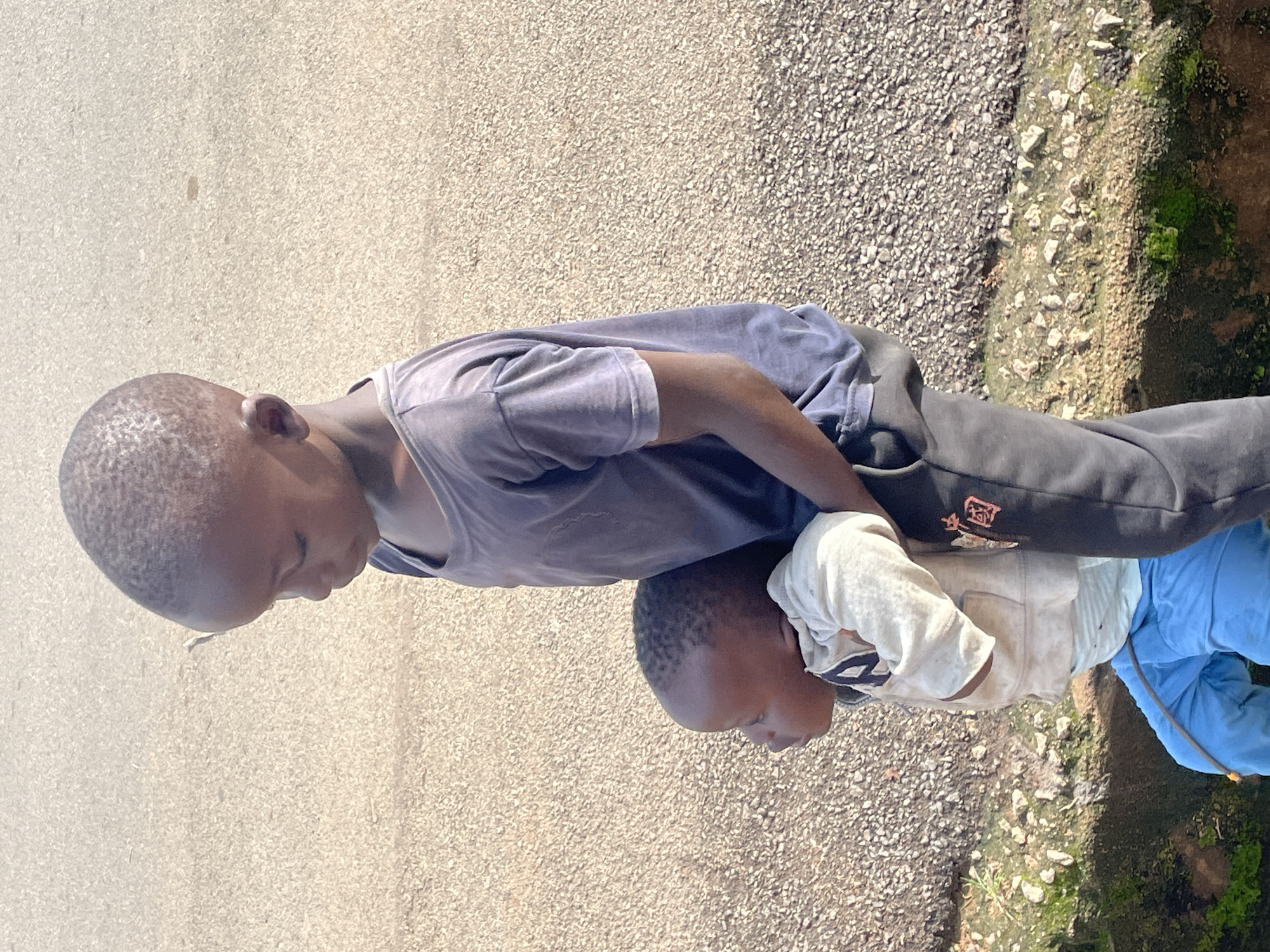In Barakin Lerawi, Nigeria, two young children are positioned on a paved surface, with one child, an older boy, standing and holding a younger child. The older boy wears a dark blue t-shirt and dark pants, and the younger child wears a light-colored hooded jacket over a white shirt, and light blue pants. The environment appears to be outdoors, with a rough, gravelly ground beside a patch of greenery.