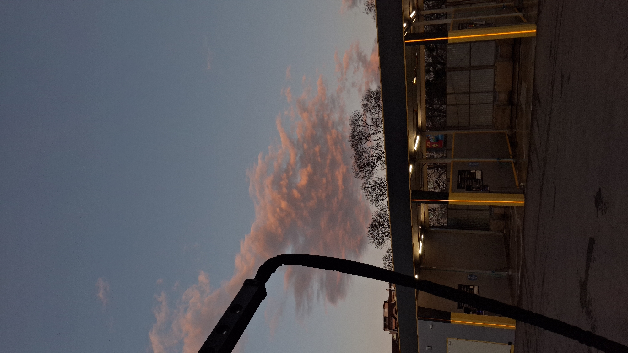 The image depicts a sideways view of an outdoor carwash at dusk. The sky is a mix of light blue and orange clouds. A black, curved structure resembling a carwash arm is in the foreground. The carwash structure has multiple bays with illuminated yellow lights and signs. Bare trees are visible behind the carwash bays. The ground is paved and dark.